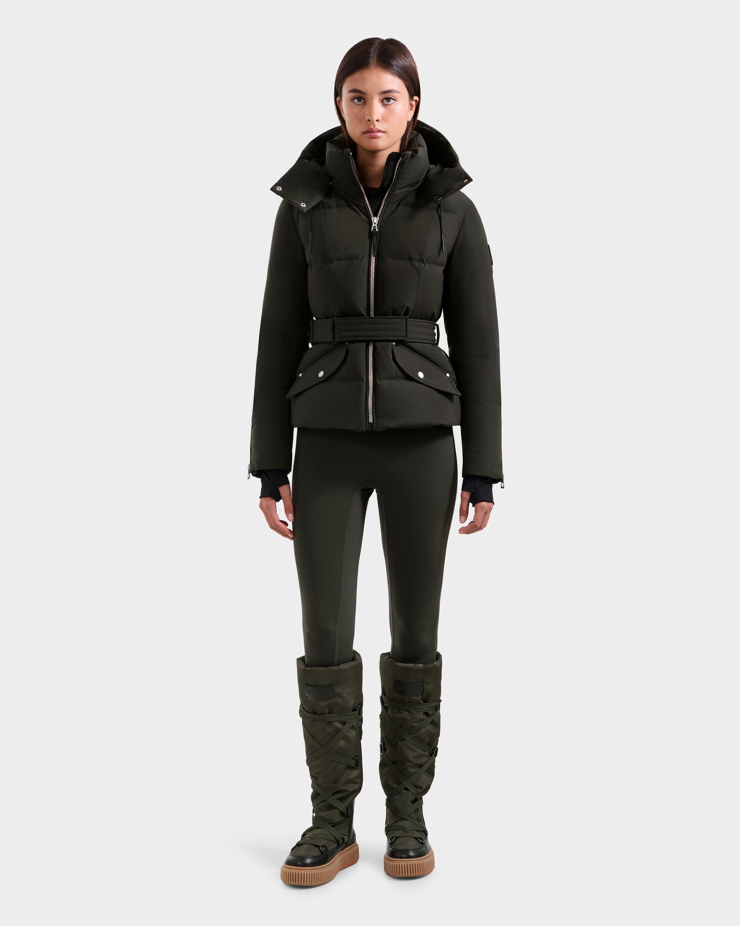A woman faces forward in a Rudsak ADELINE H - 8125587 dark olive women's down puffer jacket with a belted waist, paired with matching fitted leggings and knee-high quilted boots against a plain light gray background.