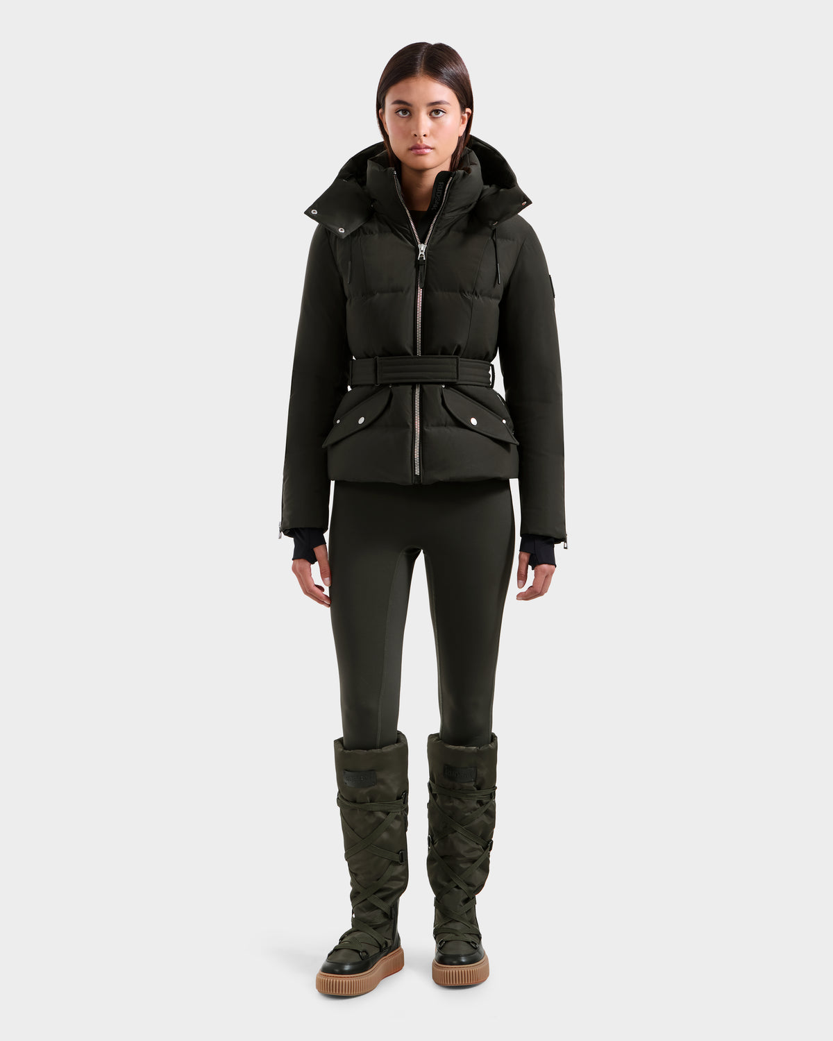 A woman faces forward in a Rudsak ADELINE H - 8125587 dark olive women's down puffer jacket with a belted waist, paired with matching fitted leggings and knee-high quilted boots against a plain light gray background.