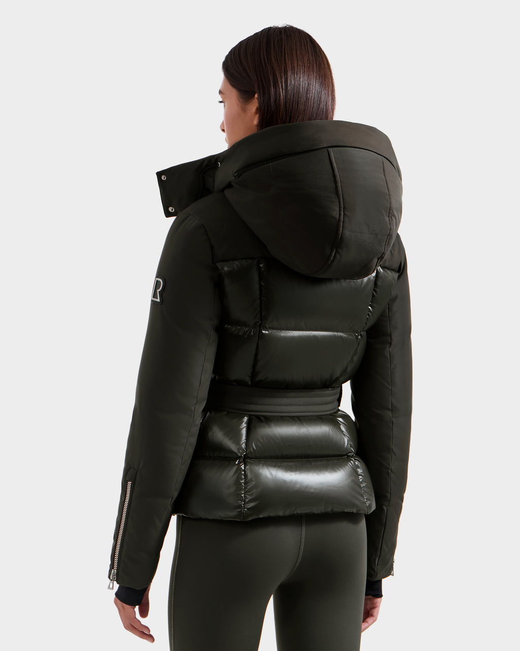 A woman with straight dark hair is seen from the back wearing the shiny, fitted Rudsak ADELINE H - 8125587 Dark Olive jacket with hood and large collar, paired with matching leggings against a plain background.