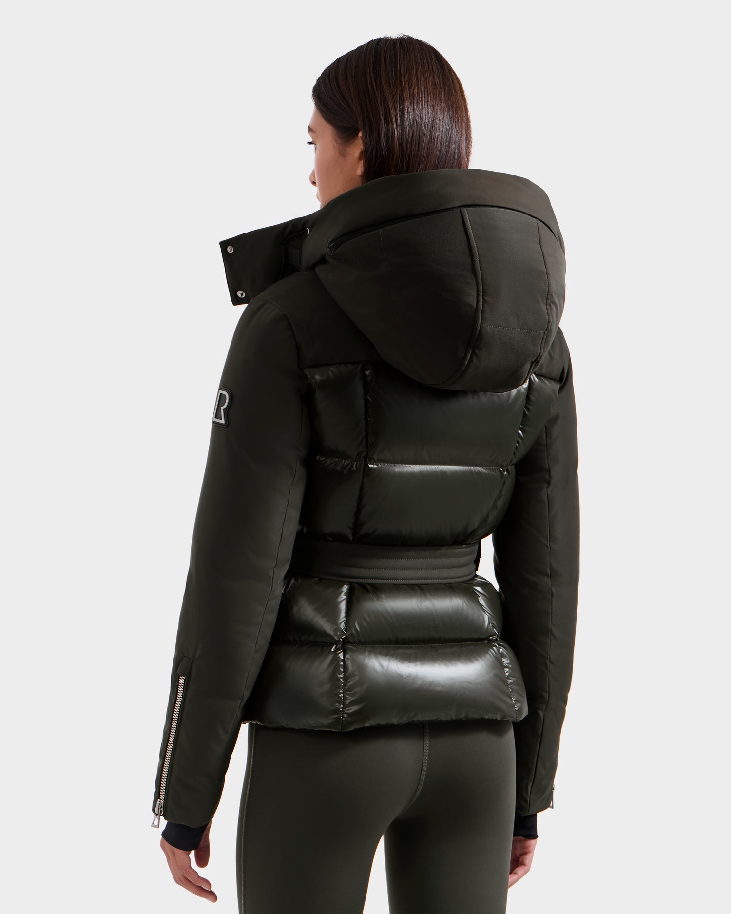 A woman with straight dark hair is seen from the back wearing the shiny, fitted Rudsak ADELINE H - 8125587 Dark Olive jacket with hood and large collar, paired with matching leggings against a plain background.