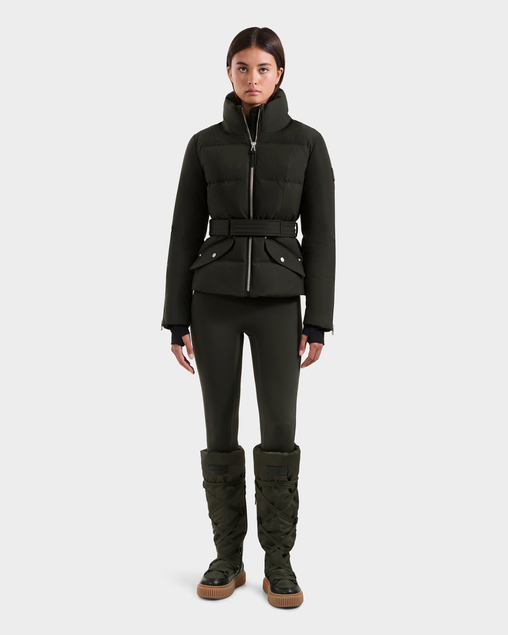 A woman stands facing forward in a dark olive ADELINE H - 8125587 down puffer jacket by Rudsak, styled with a belt, matching fitted pants, and tall padded boots against a plain white background.