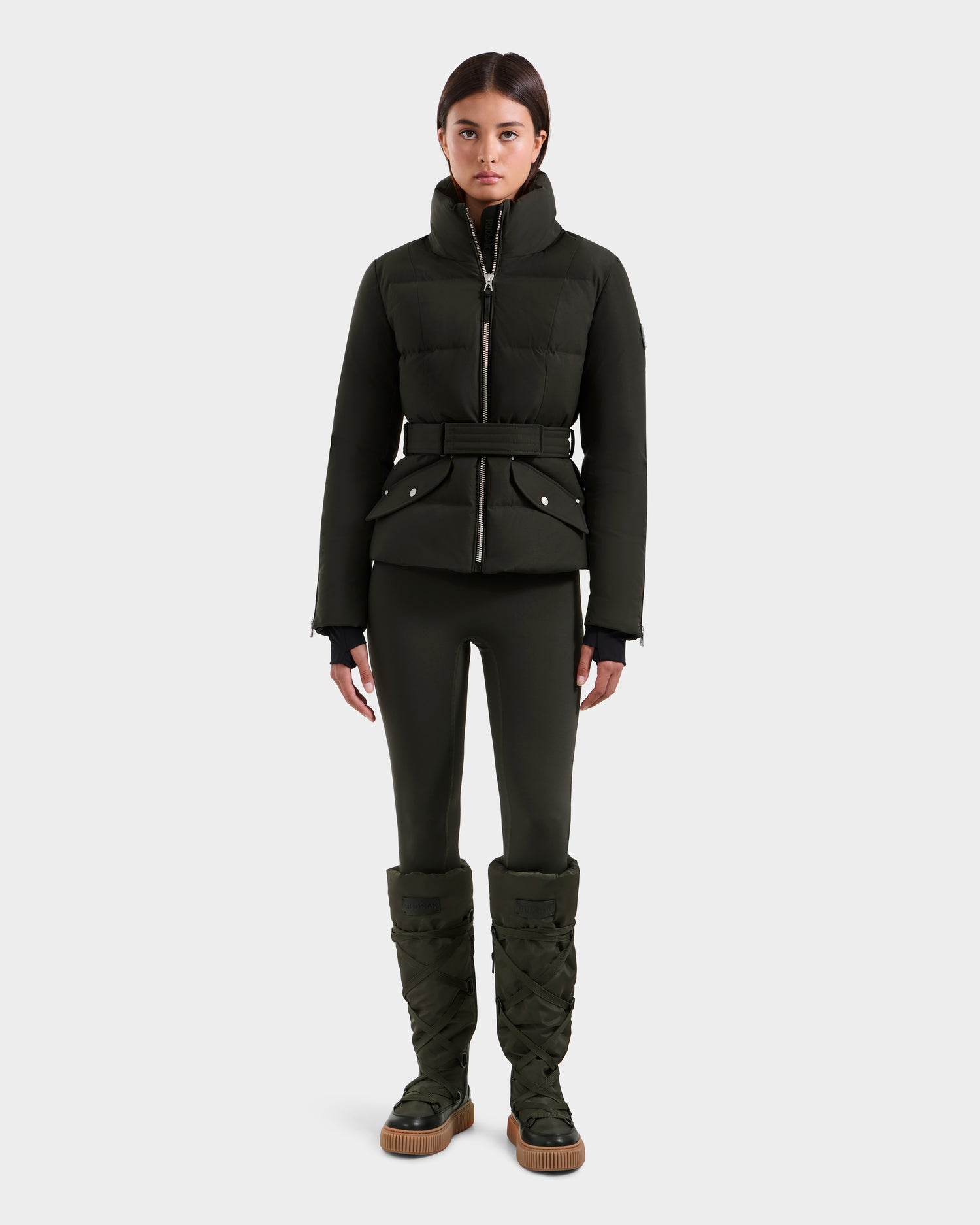A woman stands facing forward in a dark olive ADELINE H - 8125587 down puffer jacket by Rudsak, styled with a belt, matching fitted pants, and tall padded boots against a plain white background.