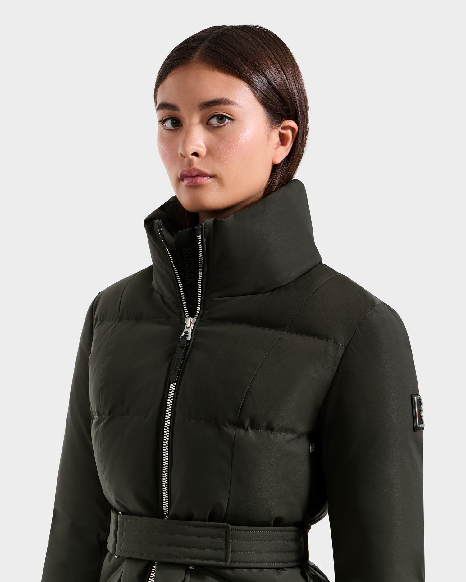 A woman with straight dark hair models the Rudsak ADELINE H - 8125587 Dark Olive jacket, featuring a high collar, front zipper, and belted waist, as she poses against a plain light background to showcase this stylish winter coat.