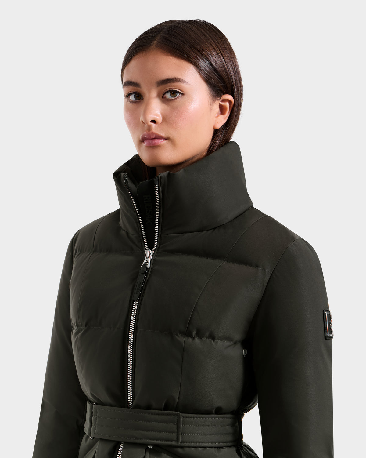 A woman with straight dark hair models the Rudsak ADELINE H - 8125587 Dark Olive jacket, featuring a high collar, front zipper, and belted waist, as she poses against a plain light background to showcase this stylish winter coat.