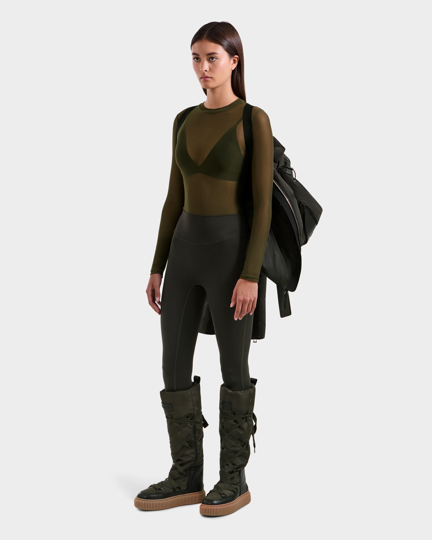 A woman stands facing forward in a sheer olive top, matching bralette, dark high-waisted leggings, large olive boots, and the Rudsak ADELINE H - 8125587 Dark Olive jacket draped over one shoulder against a plain light background.