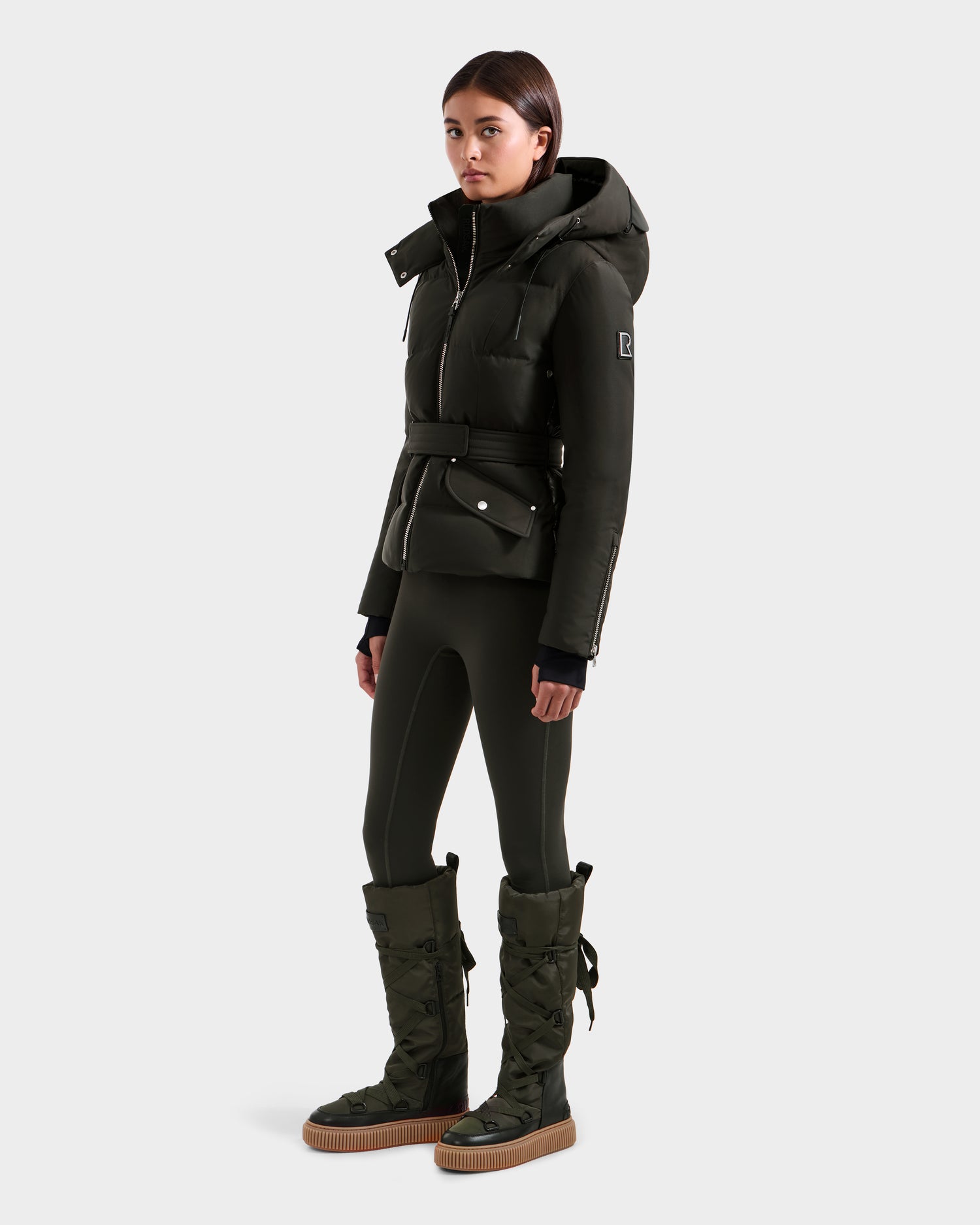 A woman faces forward in the Rudsak ADELINE H - 8125587 Dark Olive women’s down puffer winter coat with a belt, matching fitted leggings, and tall lace-up boots with gum soles against a plain white background.