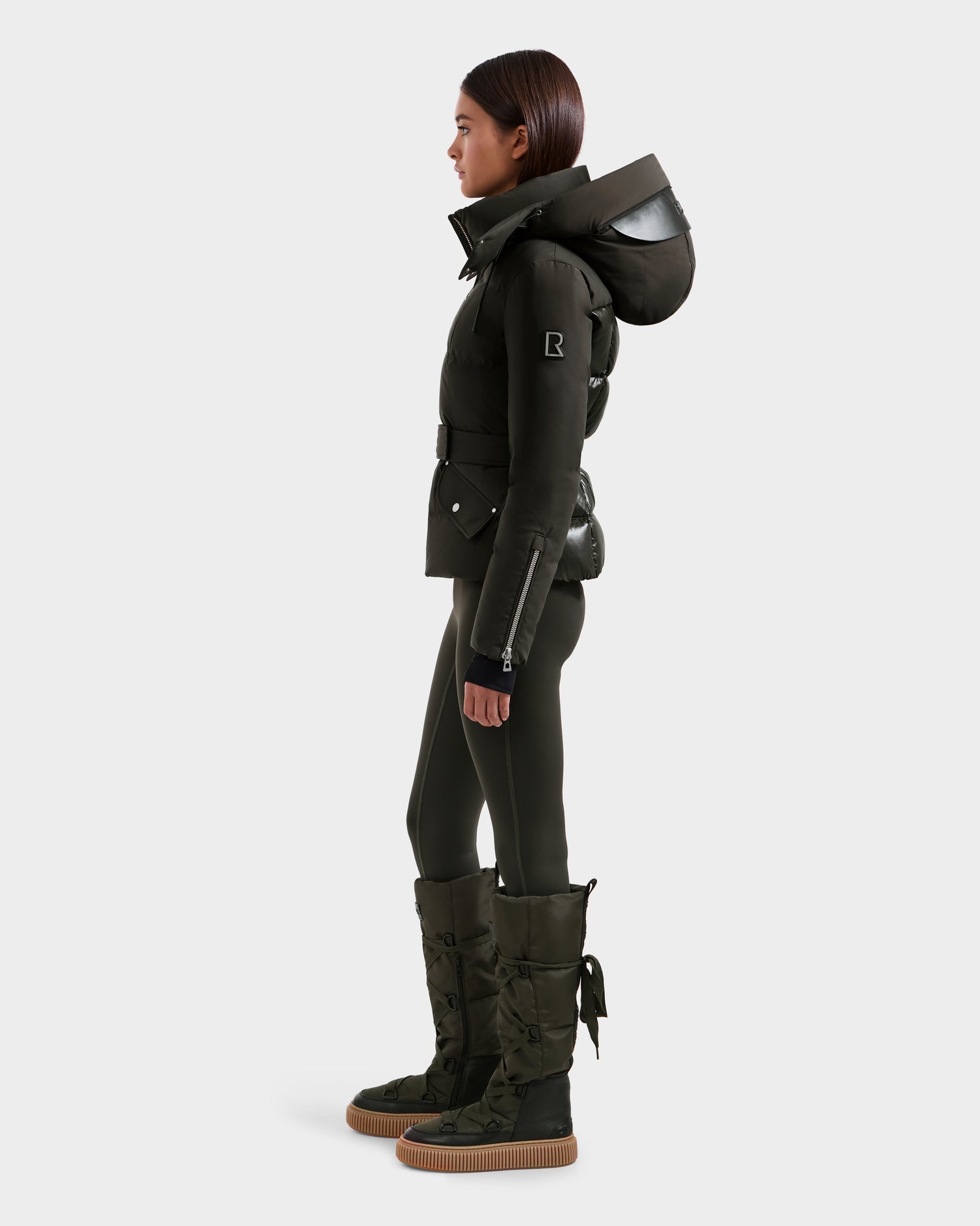 A woman poses in profile against a light gray background wearing the Rudsak ADELINE H - 8125587 Dark Olive jacket with a large hood, matching fitted pants, and tall quilted flat-soled boots.