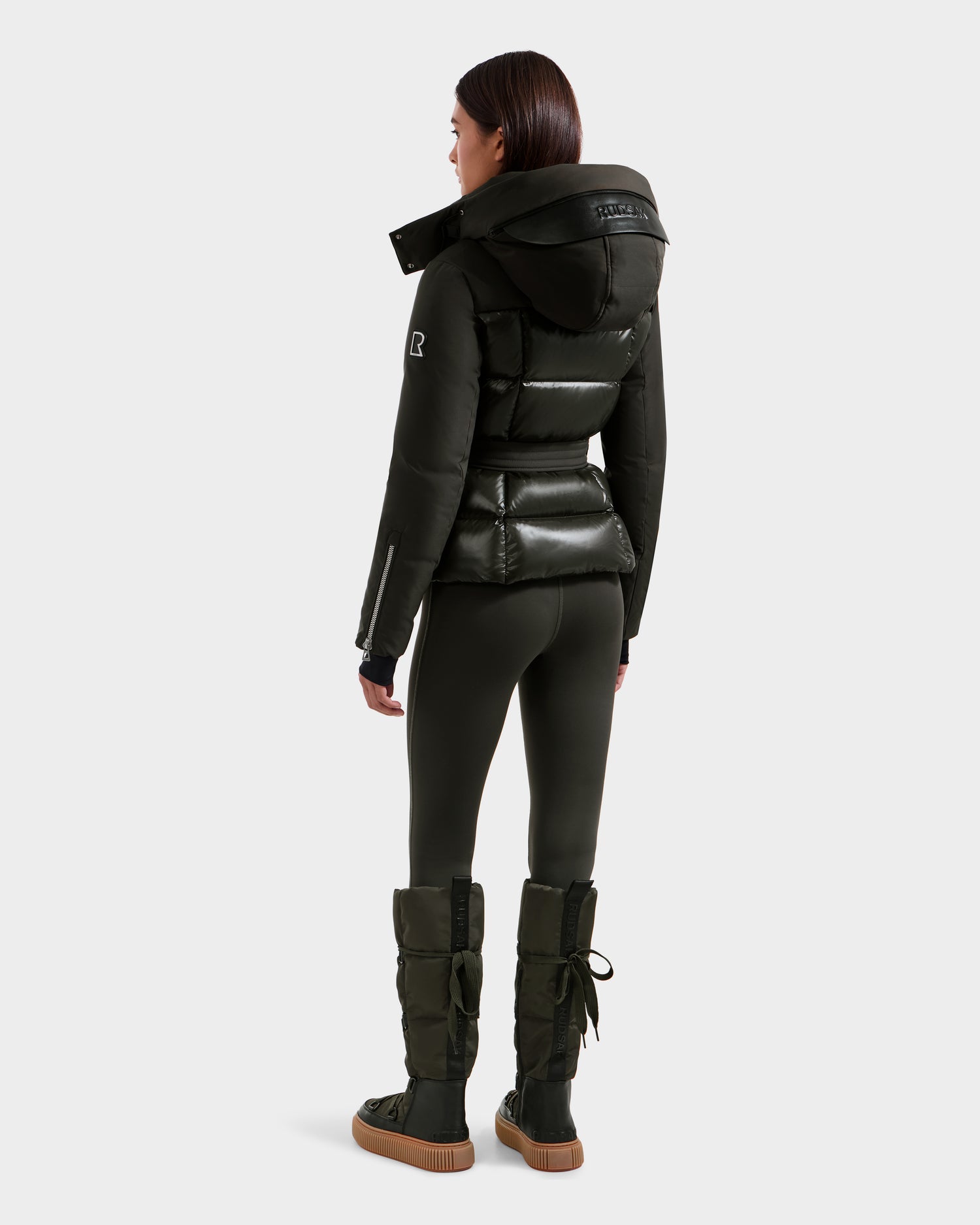 A woman faces away, wearing the Rudsak ADELINE H - 8125587 jacket in Dark Olive with a hood, paired with black leggings and puffer boots against a plain light background.