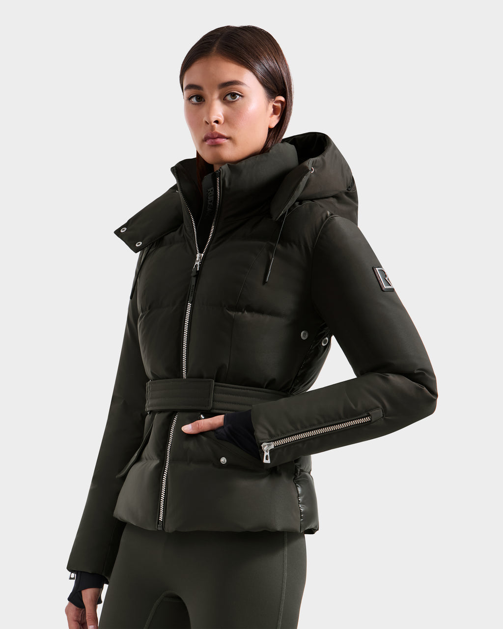 A woman with straight brown hair and a calm expression poses against a plain light background, wearing the Rudsak ADELINE H - 8125587 jacket in Dark Olive, featuring a high collar, hood, waist belt, and one hand in her pocket.