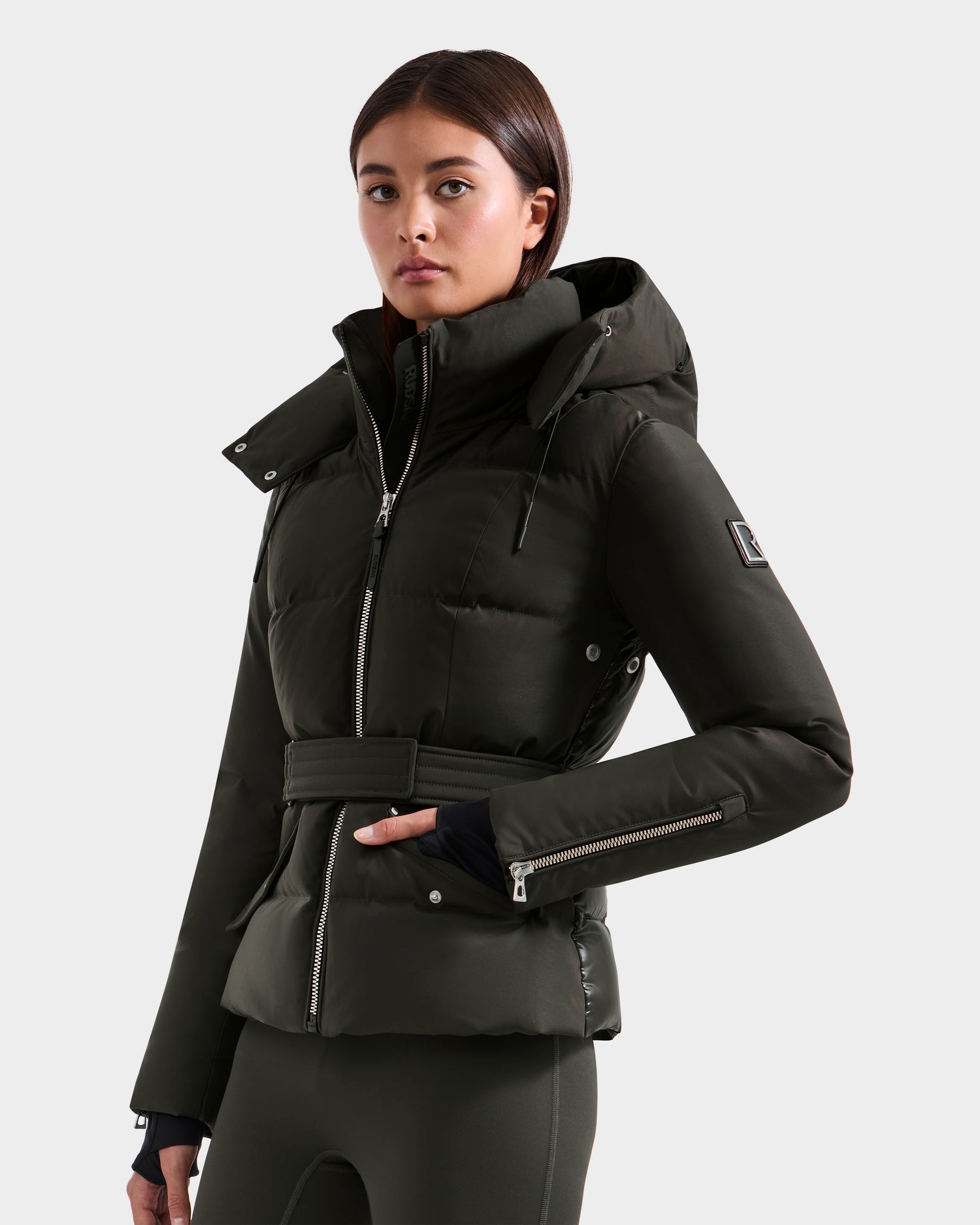 A woman with straight brown hair and a calm expression poses against a plain light background, wearing the Rudsak ADELINE H - 8125587 jacket in Dark Olive, featuring a high collar, hood, waist belt, and one hand in her pocket.