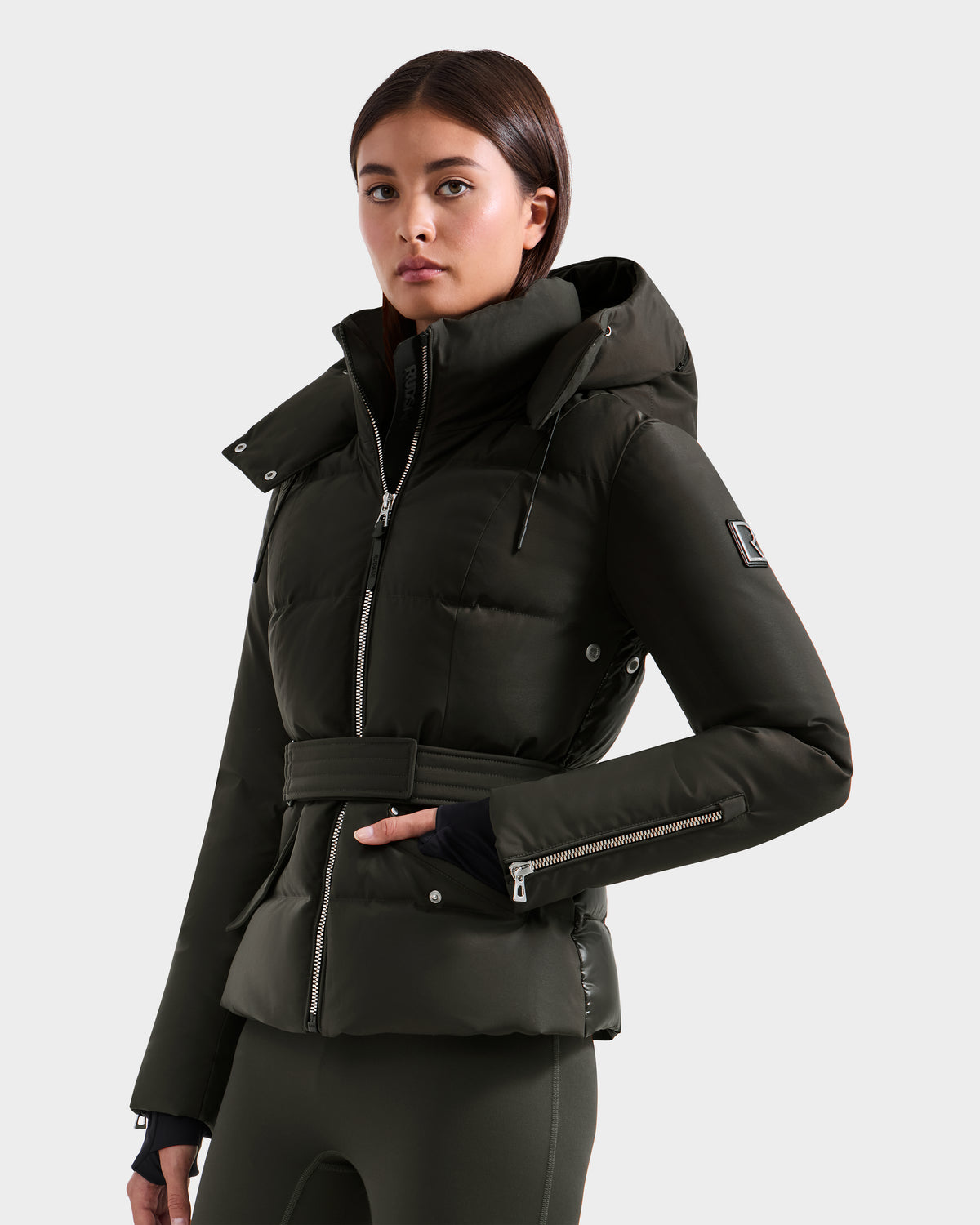 A woman with straight brown hair and a calm expression poses against a plain light background, wearing the Rudsak ADELINE H - 8125587 jacket in Dark Olive, featuring a high collar, hood, waist belt, and one hand in her pocket.