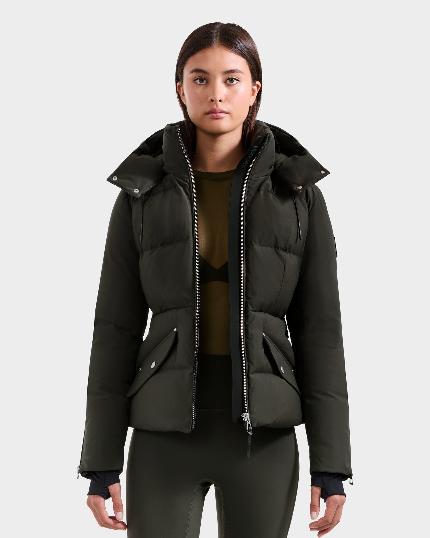 A woman stands facing forward against a light gray background, wearing the Rudsak ADELINE H - 8125587 Dark Olive women’s down puffer over a sheer olive top and matching leggings.