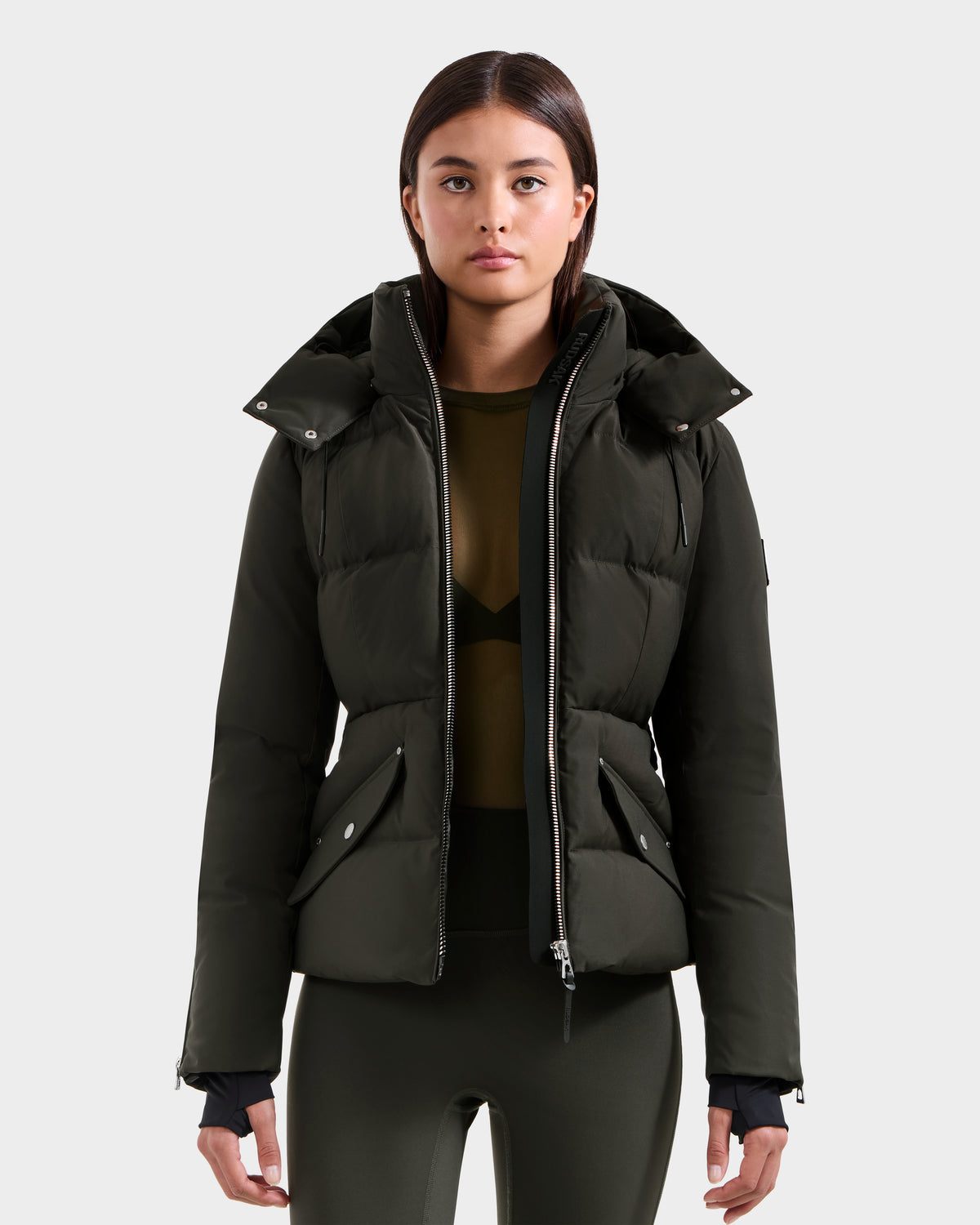A woman stands facing forward against a light gray background, wearing the Rudsak ADELINE H - 8125587 Dark Olive women’s down puffer over a sheer olive top and matching leggings.