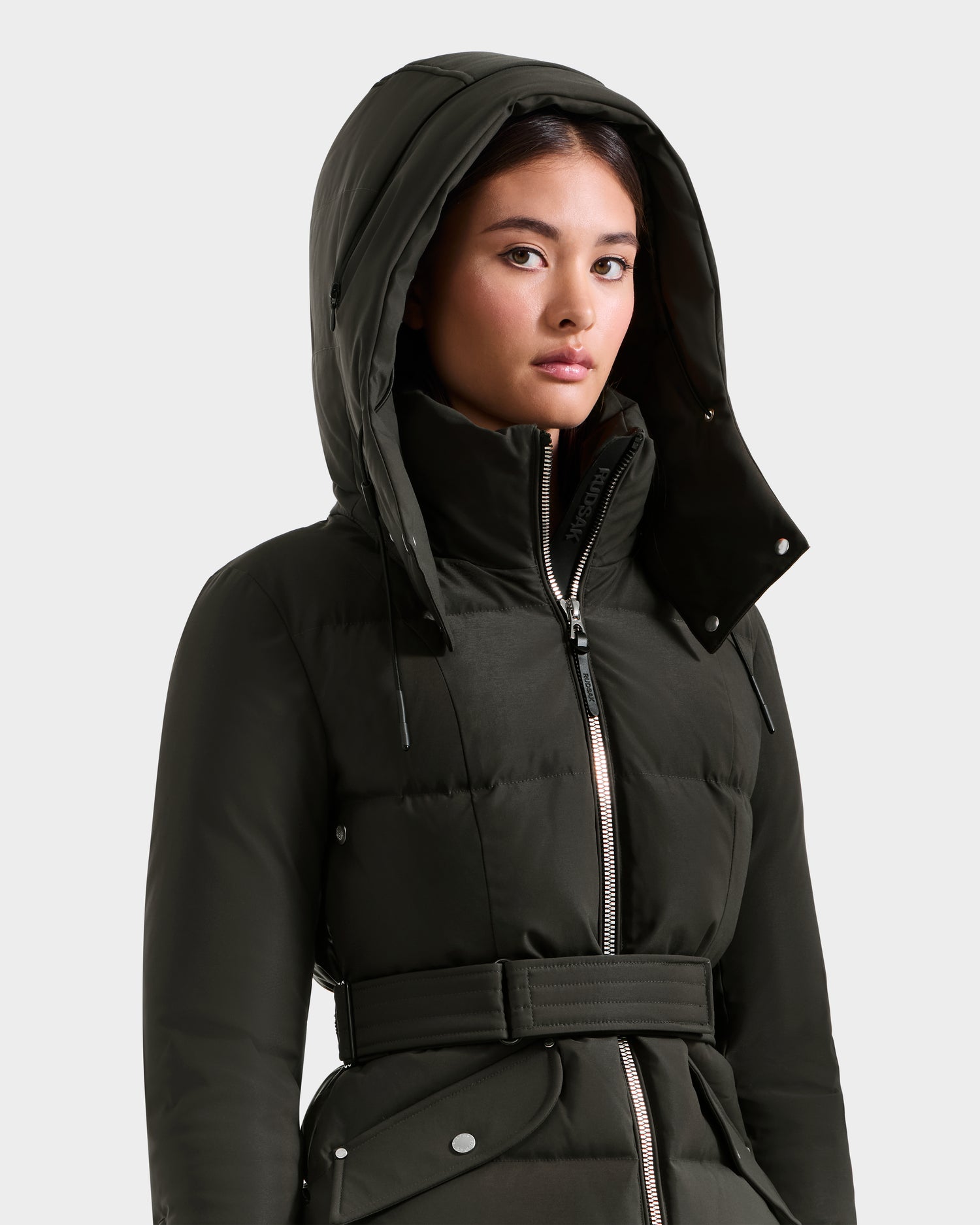 A woman in a dark olive ADELINE H - 8125587 puffer by Rudsak, with a hood up and belted waist, looks slightly to the side, highlighting this stylish women's down jacket against a plain light background.