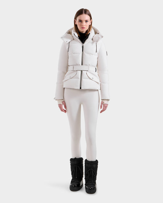 A woman faces forward in the Rudsak ADELINE H - 8125587 Vanilla, a white belted women's down puffer, styled with white fitted pants and black snow boots against a plain white background.