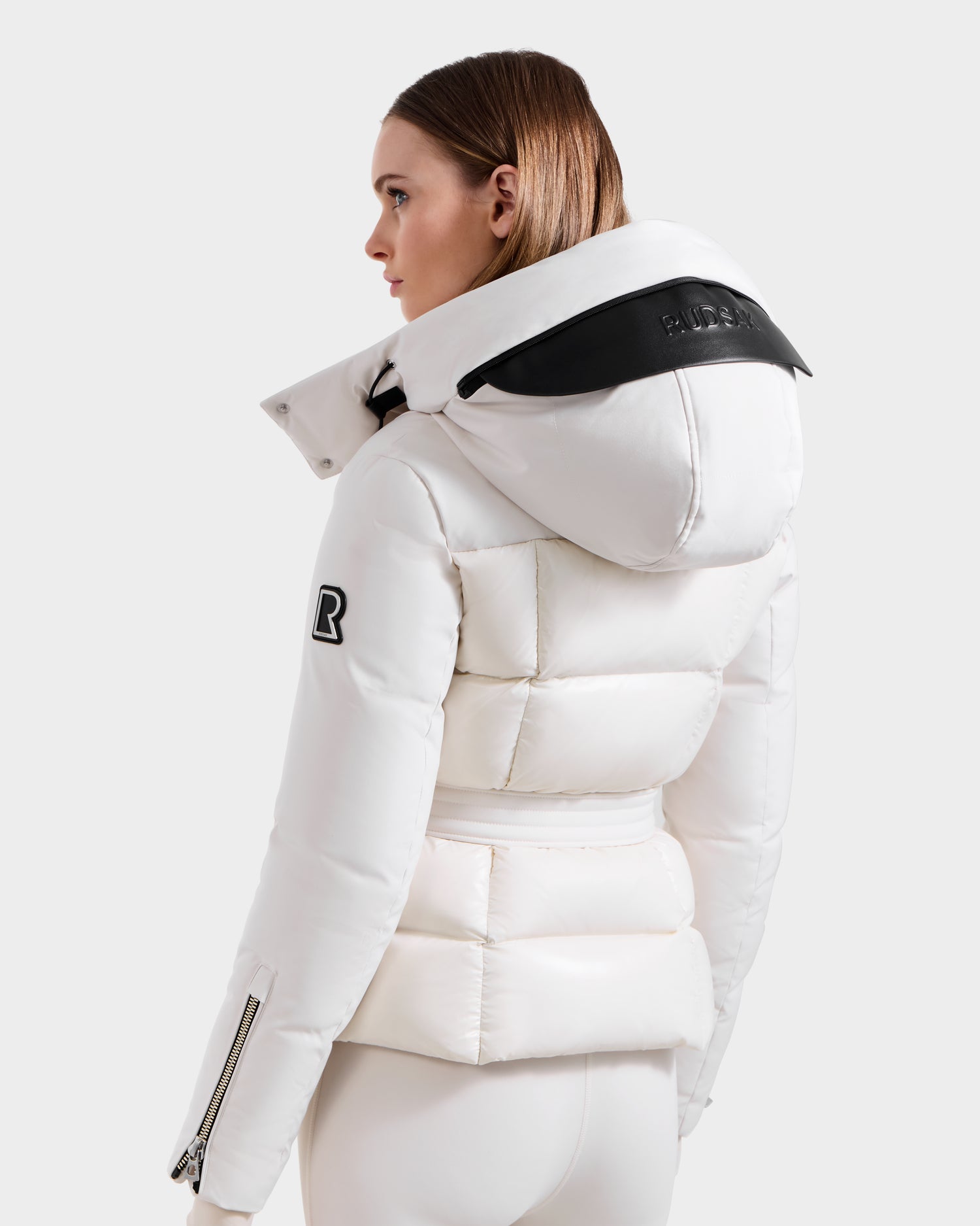 A woman with straight brown hair wears the Rudsak ADELINE H - 8125587 Vanilla down puffer, featuring a large hood, black accents, and a sleeve patch. She faces away to the left against a plain light background.