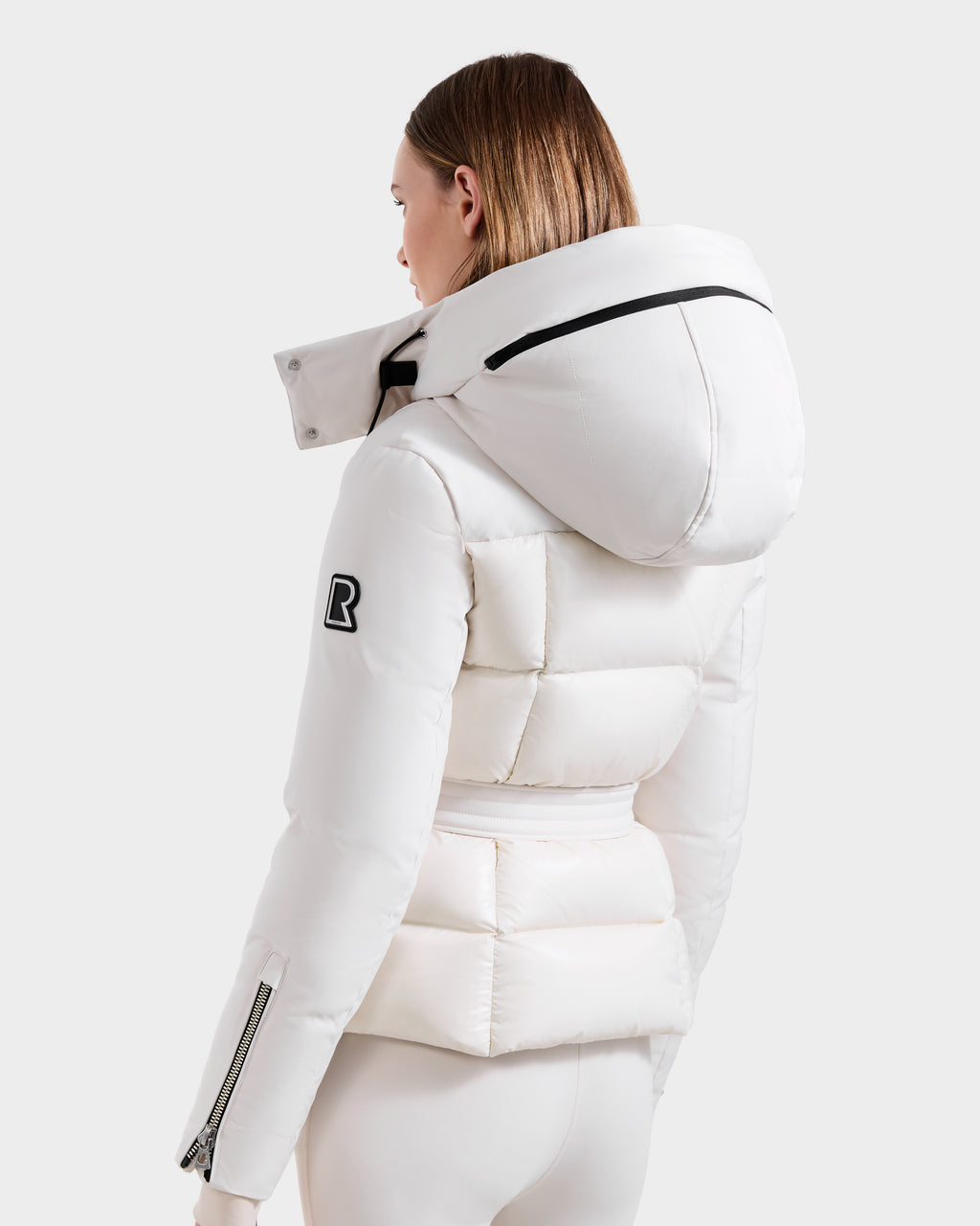 A person is seen from behind wearing the vanilla ADELINE H - 8125587 down puffer by Rudsak. This women’s winter jacket features a hood, black zipper details, and a black R patch on the left sleeve, set against a light gray background.