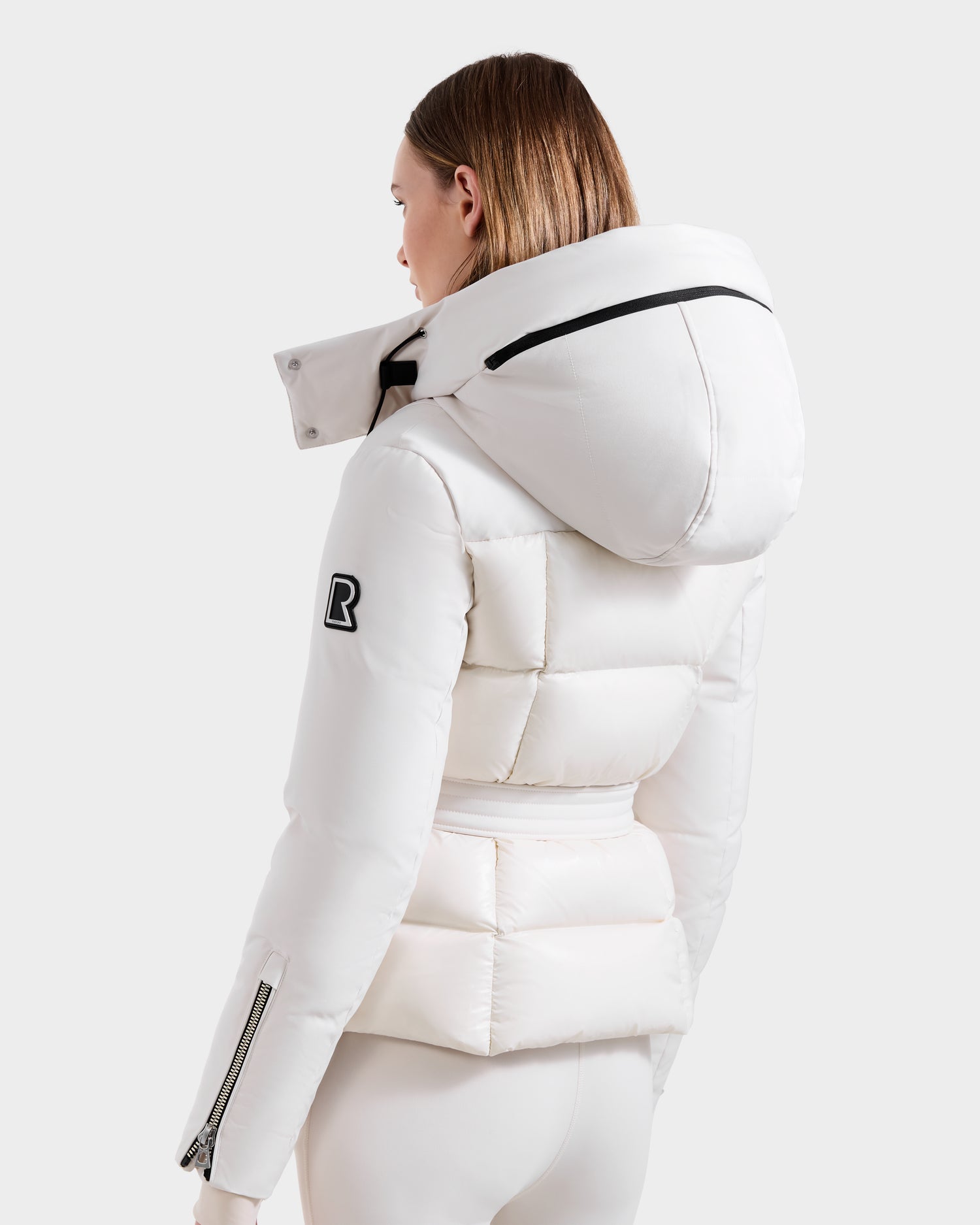 A person is seen from behind wearing the vanilla ADELINE H - 8125587 down puffer by Rudsak. This women’s winter jacket features a hood, black zipper details, and a black R patch on the left sleeve, set against a light gray background.