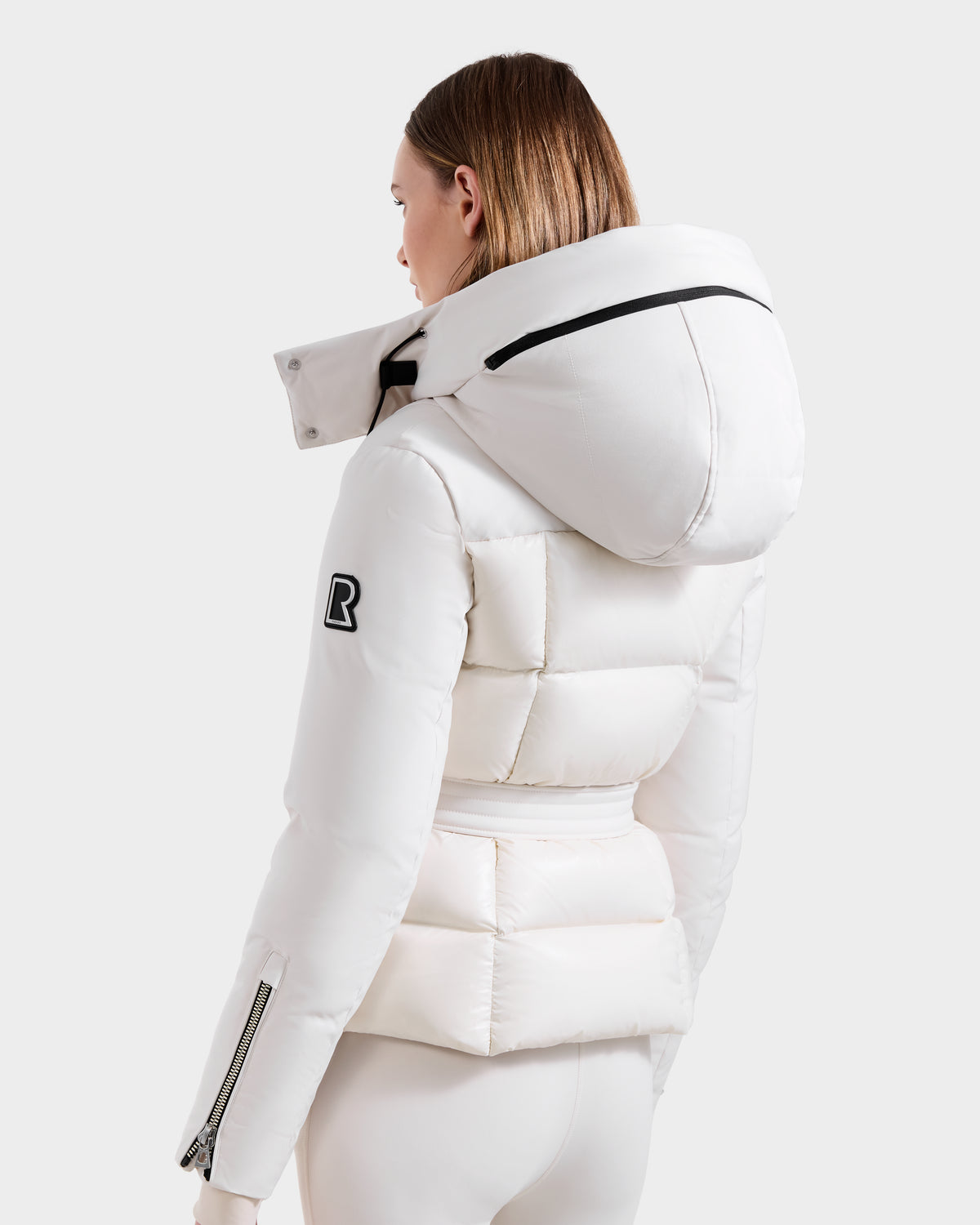 A person is seen from behind wearing the vanilla ADELINE H - 8125587 down puffer by Rudsak. This women’s winter jacket features a hood, black zipper details, and a black R patch on the left sleeve, set against a light gray background.
