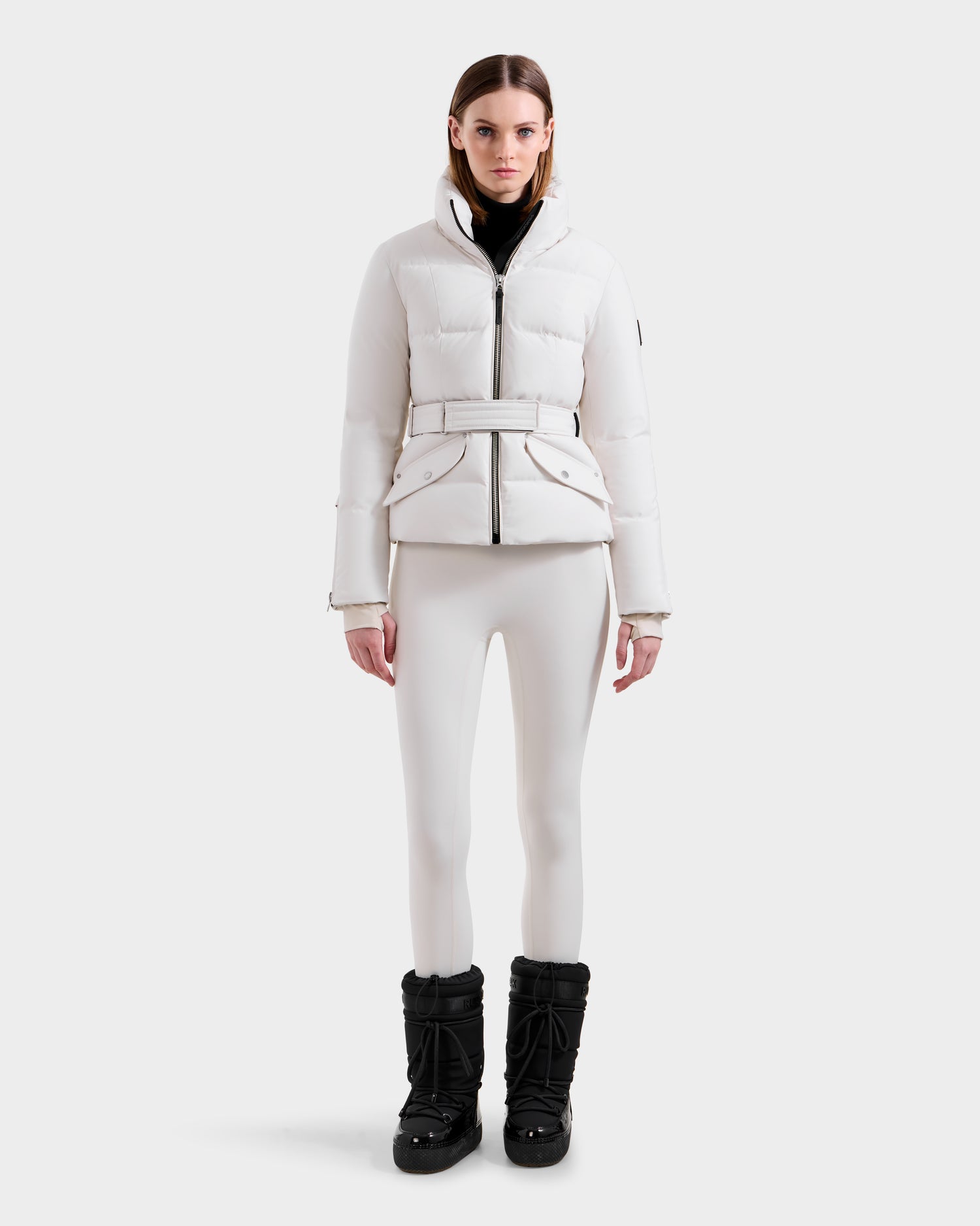 A woman models the Rudsak ADELINE H - 8125587 Vanilla down puffer jacket against a white background, paired with fitted pants and a black turtleneck, highlighting this chic winter look.