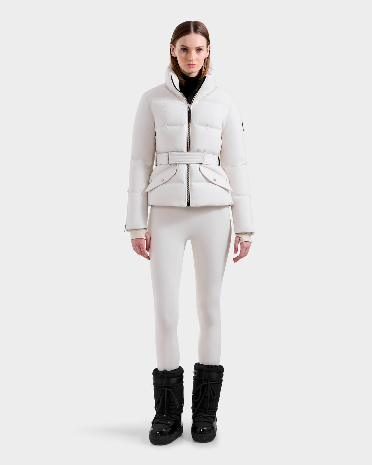 A woman models the Rudsak ADELINE H - 8125587 Vanilla down puffer jacket against a white background, paired with fitted pants and a black turtleneck, highlighting this chic winter look.