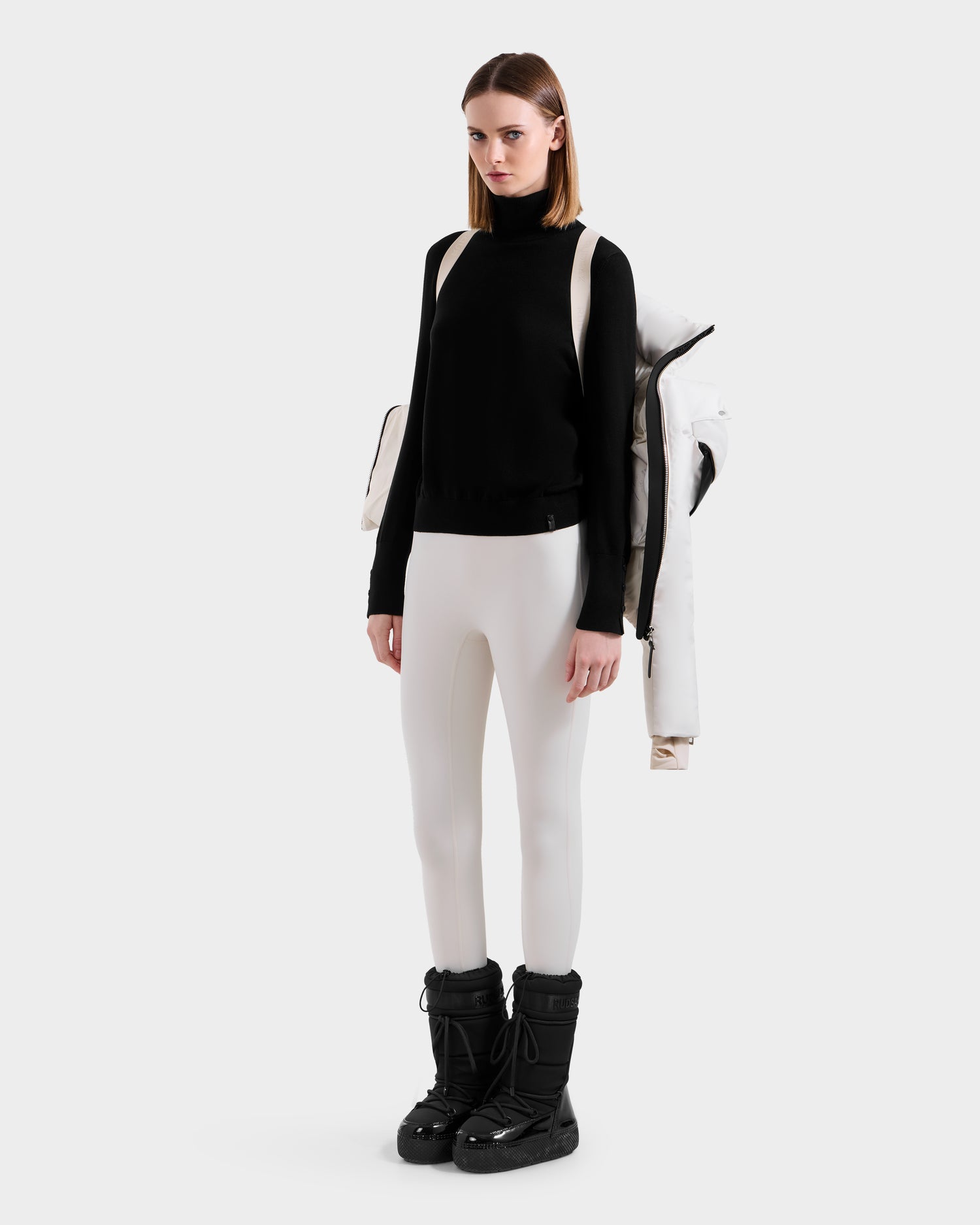 A woman faces forward in a black turtleneck, white fitted pants, and black snow boots. The ADELINE H - 8125587 Vanilla Rudsak women’s down puffer jacket is draped over her shoulders like a backpack against a plain light gray background.