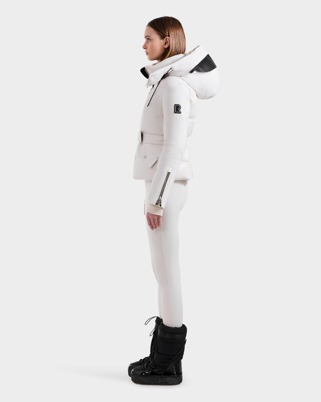 A woman stands in profile wearing the Rudsak ADELINE H - 8125587 Vanilla down puffer jacket with a large hood, paired with white pants and black snow boots. This chic winter coat offers both style and warmth against a plain white background.
