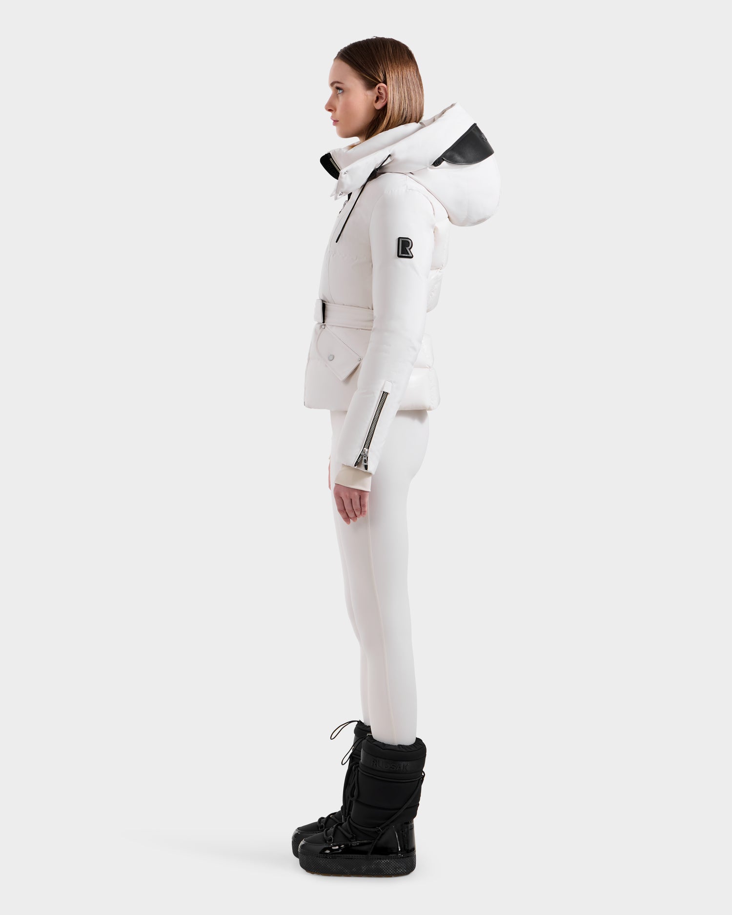 A woman stands in profile wearing the Rudsak ADELINE H - 8125587 Vanilla down puffer jacket with a large hood, paired with white pants and black snow boots. This chic winter coat offers both style and warmth against a plain white background.