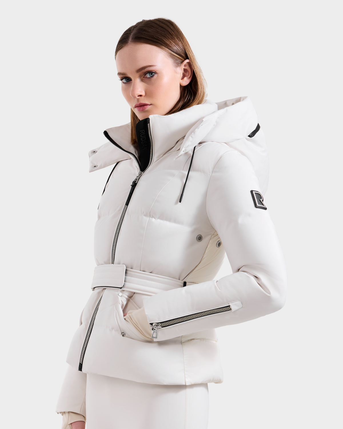 A woman wears the Rudsak ADELINE H - 8125587 Vanilla, a stylish white down puffer with a belted waist, high collar, hood, zippered pockets, and a logo patch on the sleeve, set against a plain light background.
