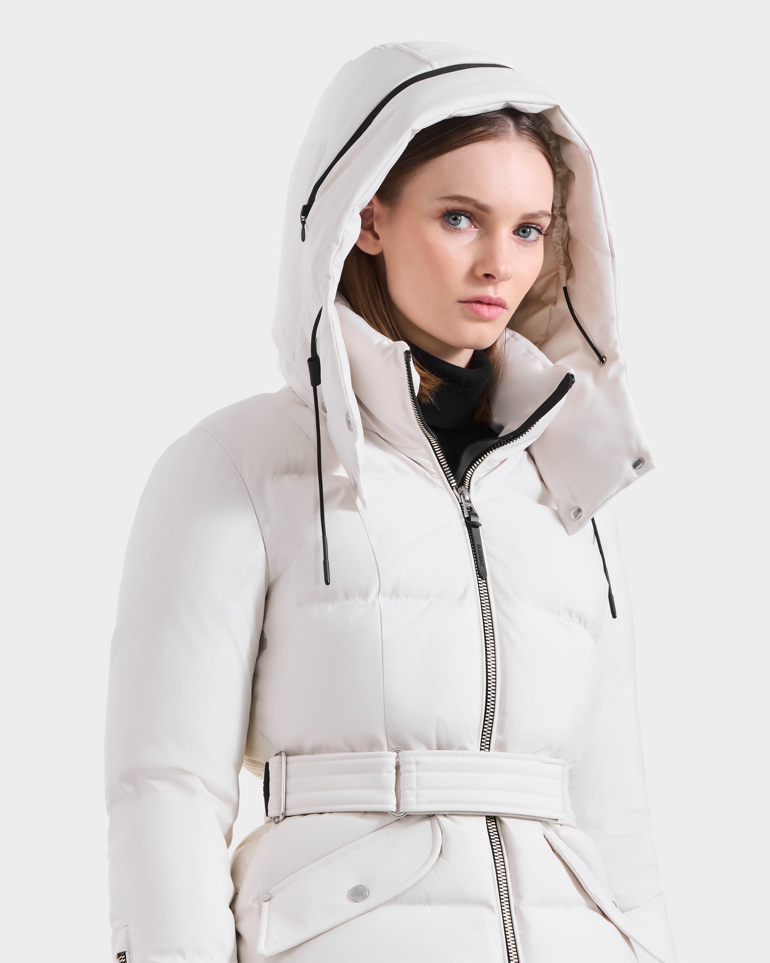 A woman in the Rudsak ADELINE H - 8125587 Vanilla, a white belted women's down puffer with a large hood and black drawstrings, stands against a plain light background, looking forward with a neutral expression.