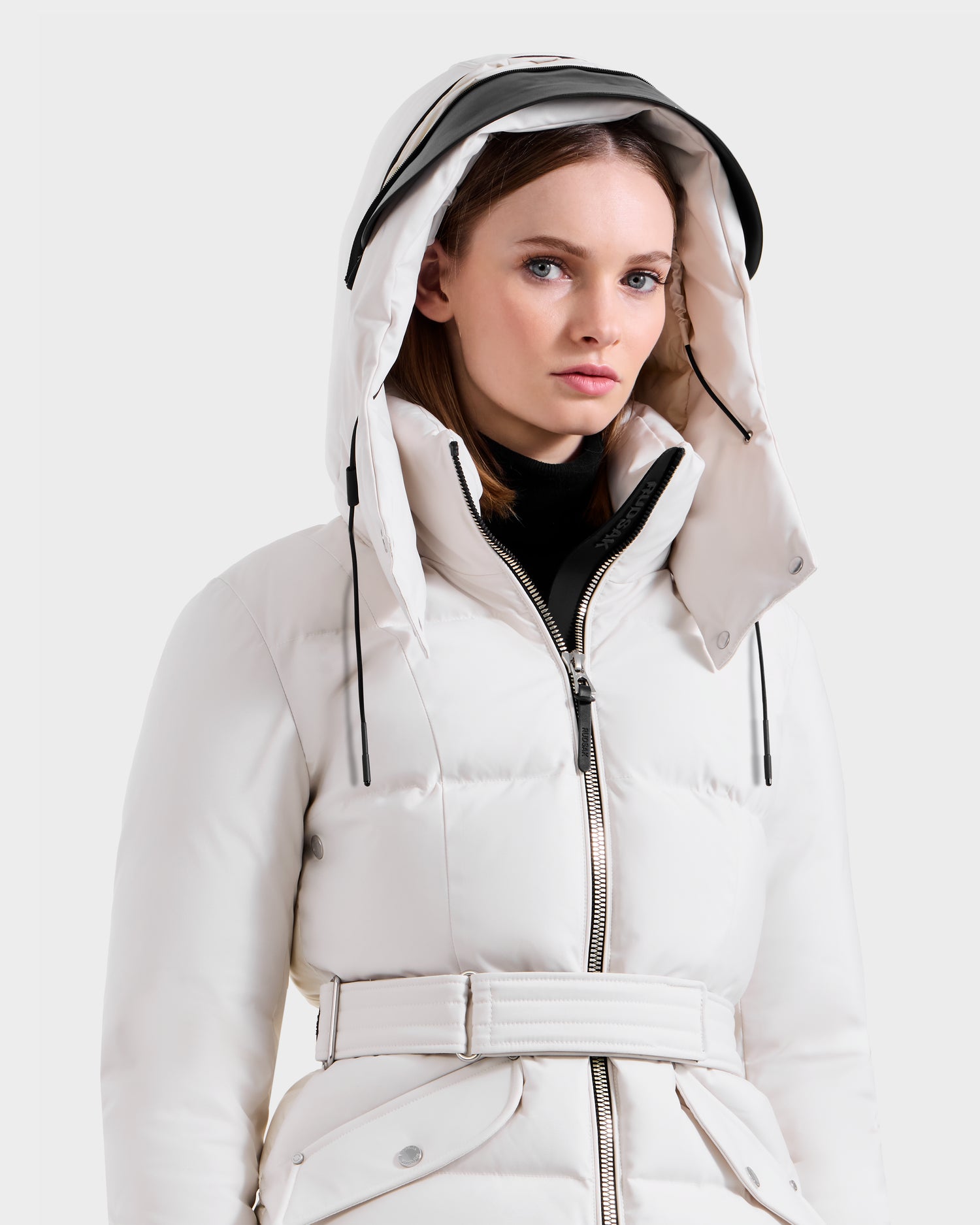 A woman wears the vanilla Rudsak ADELINE H - 8125587 down puffer with a belt, hood, and black zipper, standing against a plain light background and looking forward with a neutral expression.