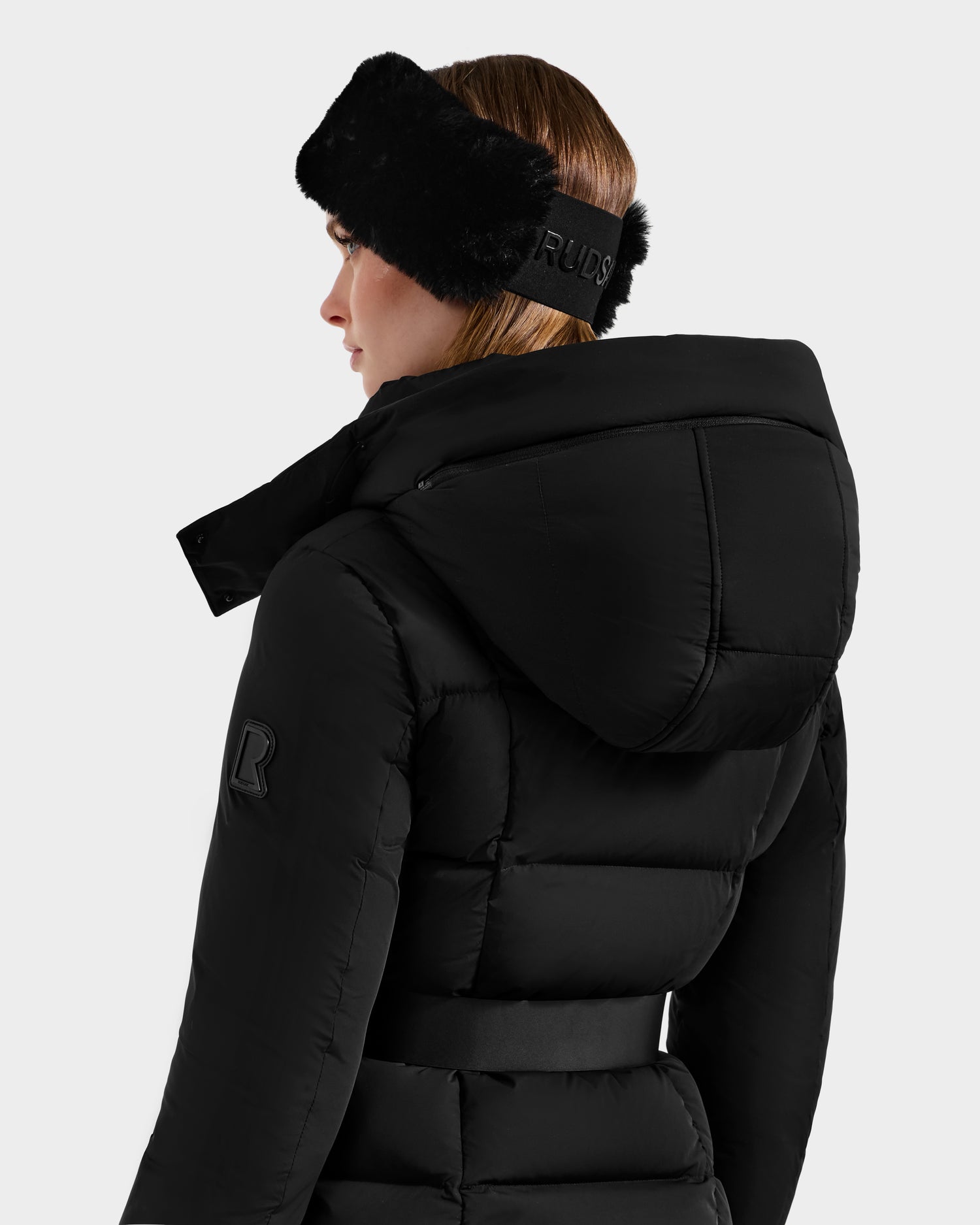 A woman with straight, light brown hair is shown from the back wearing the Rudsak ADELINE - 8124544 Black women's down puffer jacket with belted waist, large hood, R logo on the sleeve, and a black fur headband.