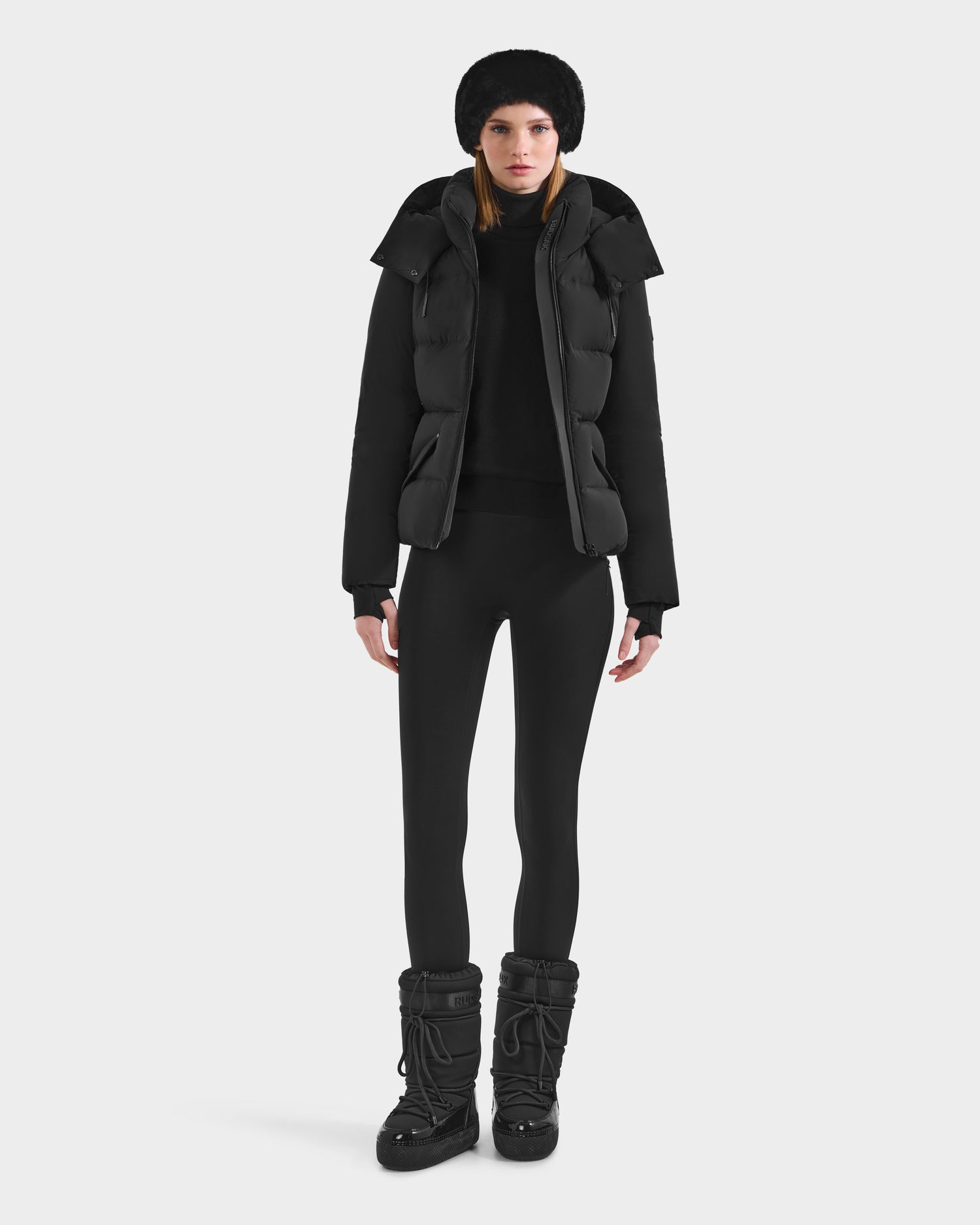 A woman faces forward against a plain white background, wearing the Rudsak ADELINE - 8124544 Black women's down puffer, paired with black leggings and black snow boots.
