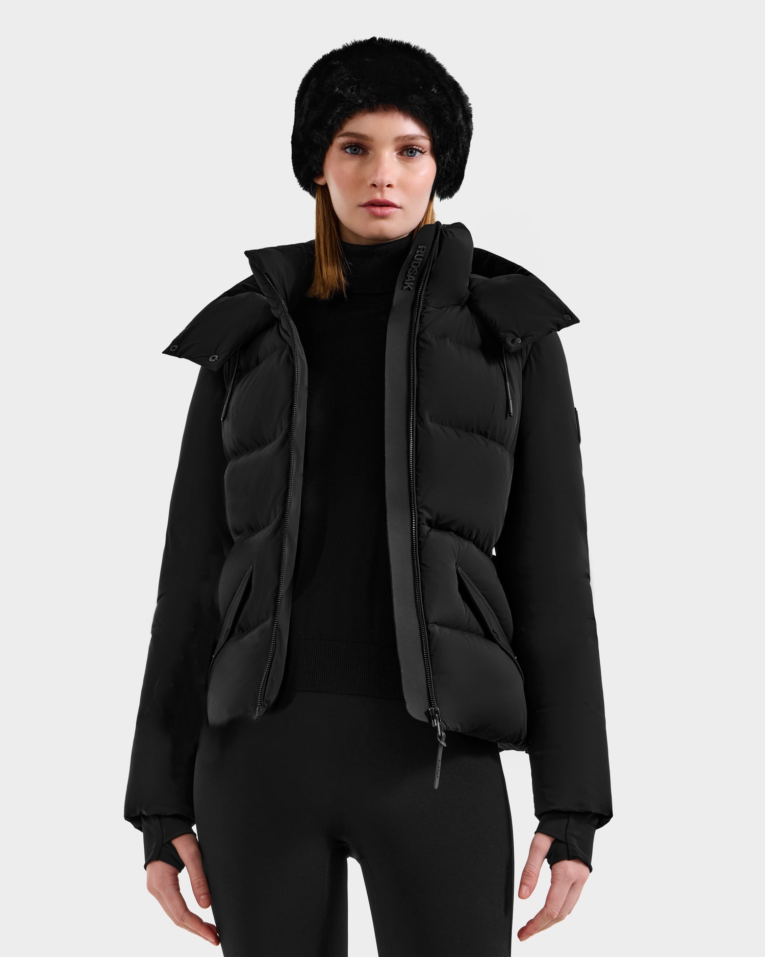 A woman in a Rudsak ADELINE - 8124544 Black down puffer, black turtleneck, black leggings, and a black fur hat stands facing forward against a plain light gray background.