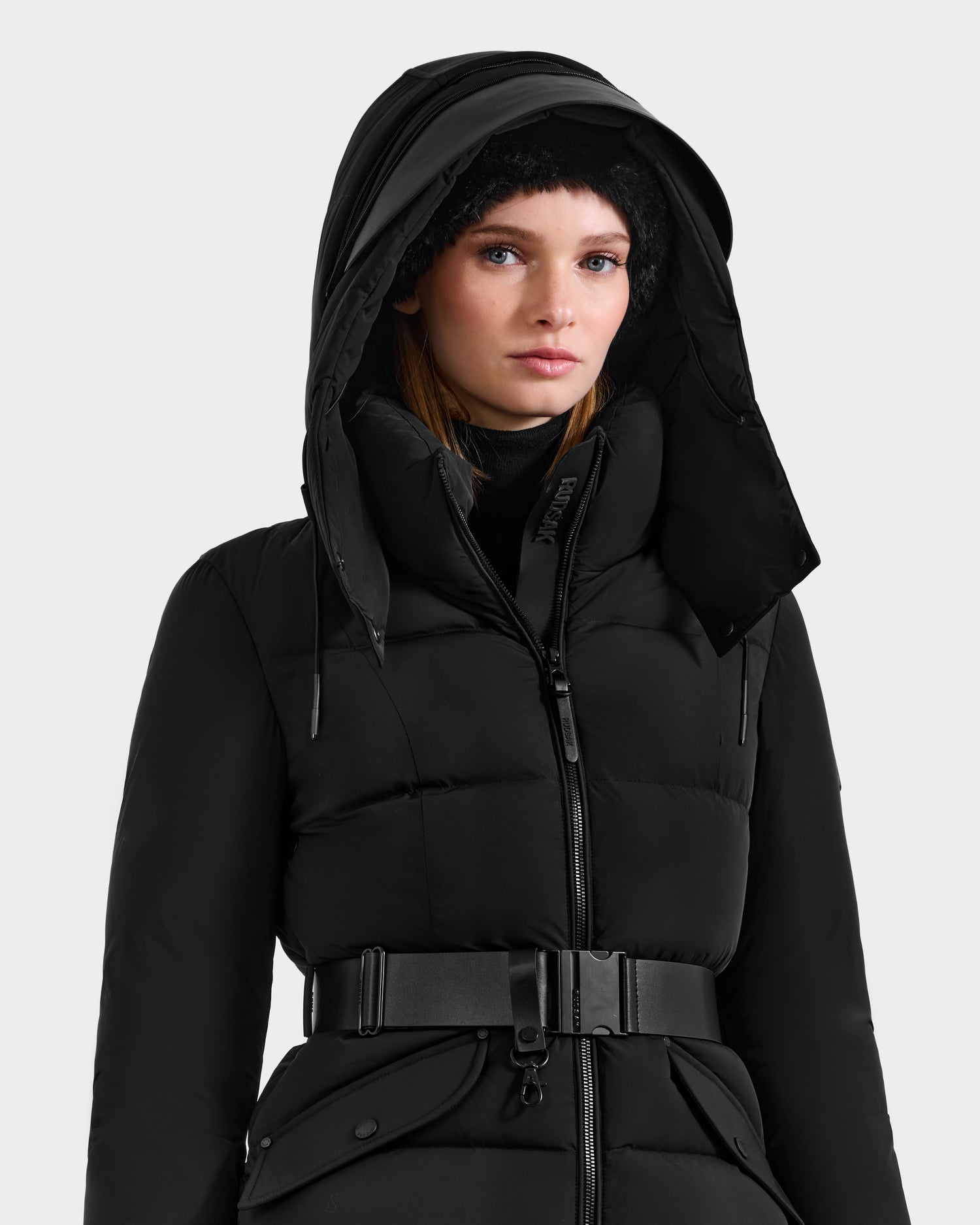 A woman models the Rudsak ADELINE - 8124544 Black quilted winter jacket with a high collar, hood, and wide belt, styled with a black beanie against a plain light background.