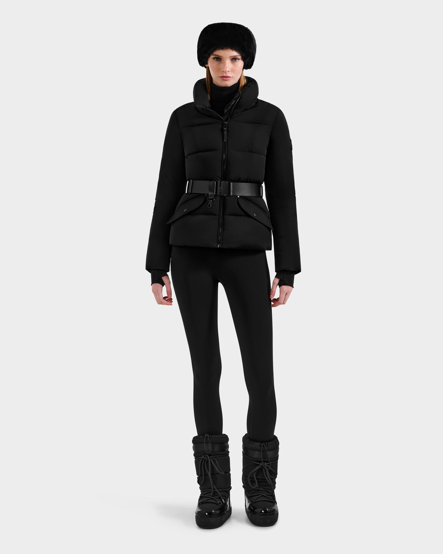 A person stands facing forward, wearing the Rudsak ADELINE - 8124544 Black quilted winter jacket with a belt, black leggings, black winter boots, and a black fur hat against a plain light background.