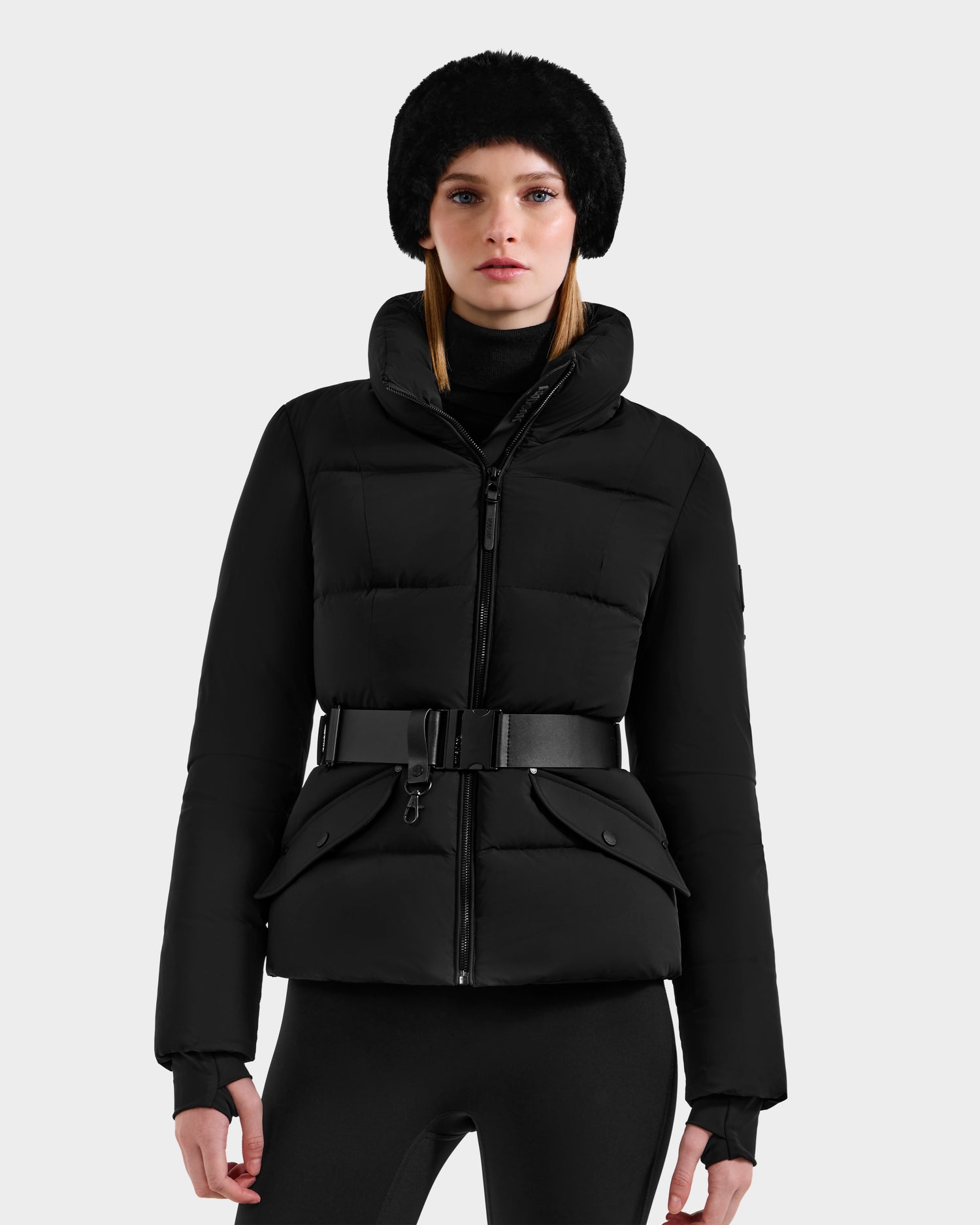A woman wearing the Rudsak ADELINE - 8124544 Black quilted winter jacket, paired with black leggings, a black turtleneck, and a furry black hat, stands against a plain light gray background.