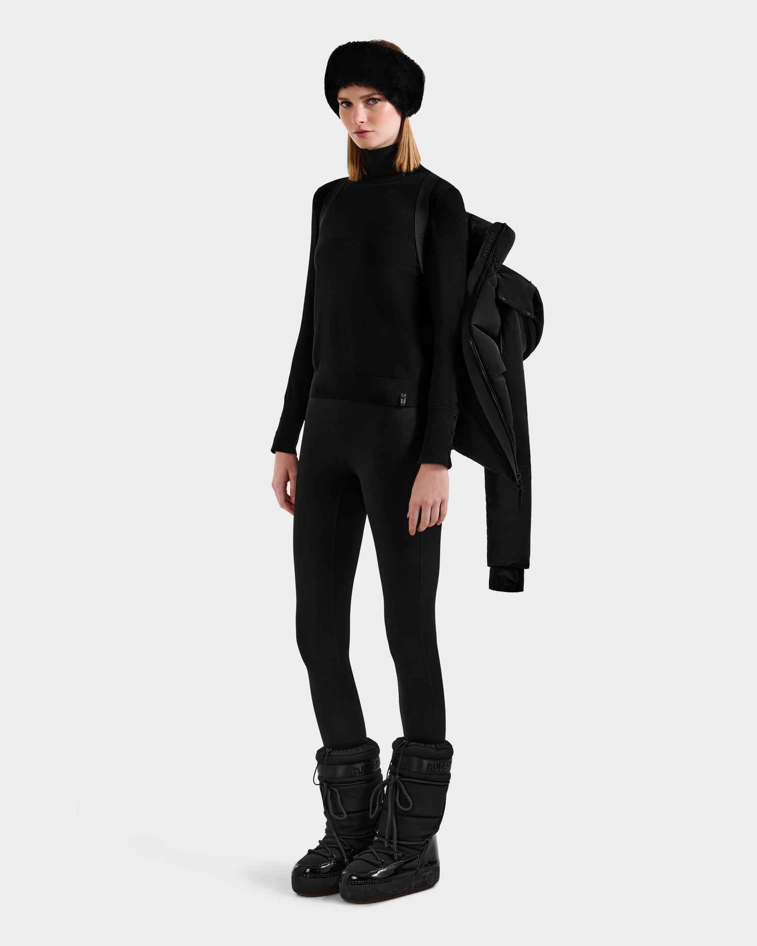 A woman poses against a plain background in an all-black look, featuring the Rudsak ADELINE - 8124544 Black women’s down puffer jacket draped over one shoulder, paired with a knit hat, sweater, leggings, and black winter boots.
