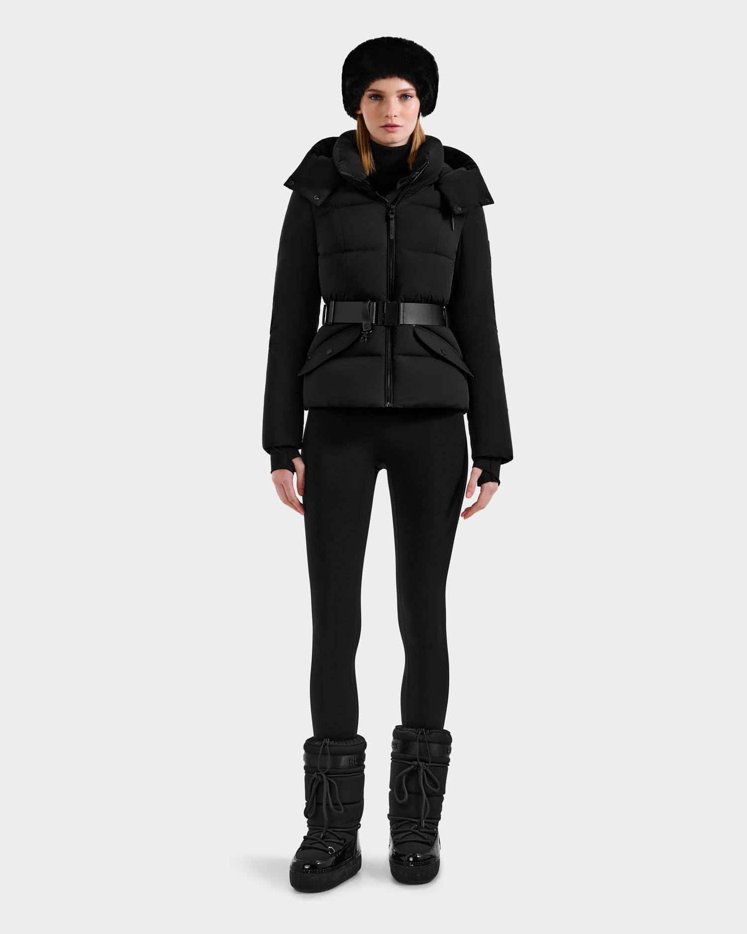 A woman faces forward wearing the Rudsak ADELINE - 8124544 Black down puffer jacket with a belt, paired with black leggings, winter boots, and a black fur hat, set against a plain light gray background.