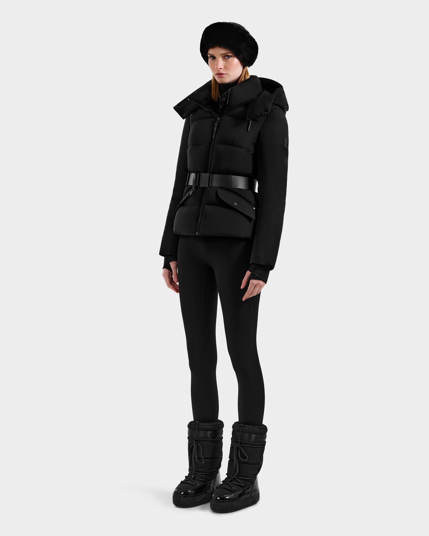 A woman stands against a plain background wearing the Rudsak ADELINE - 8124544 Black quilted winter jacket. She pairs it with fitted black pants, a black fur hat, and black winter boots, arms by her side and a neutral expression.
