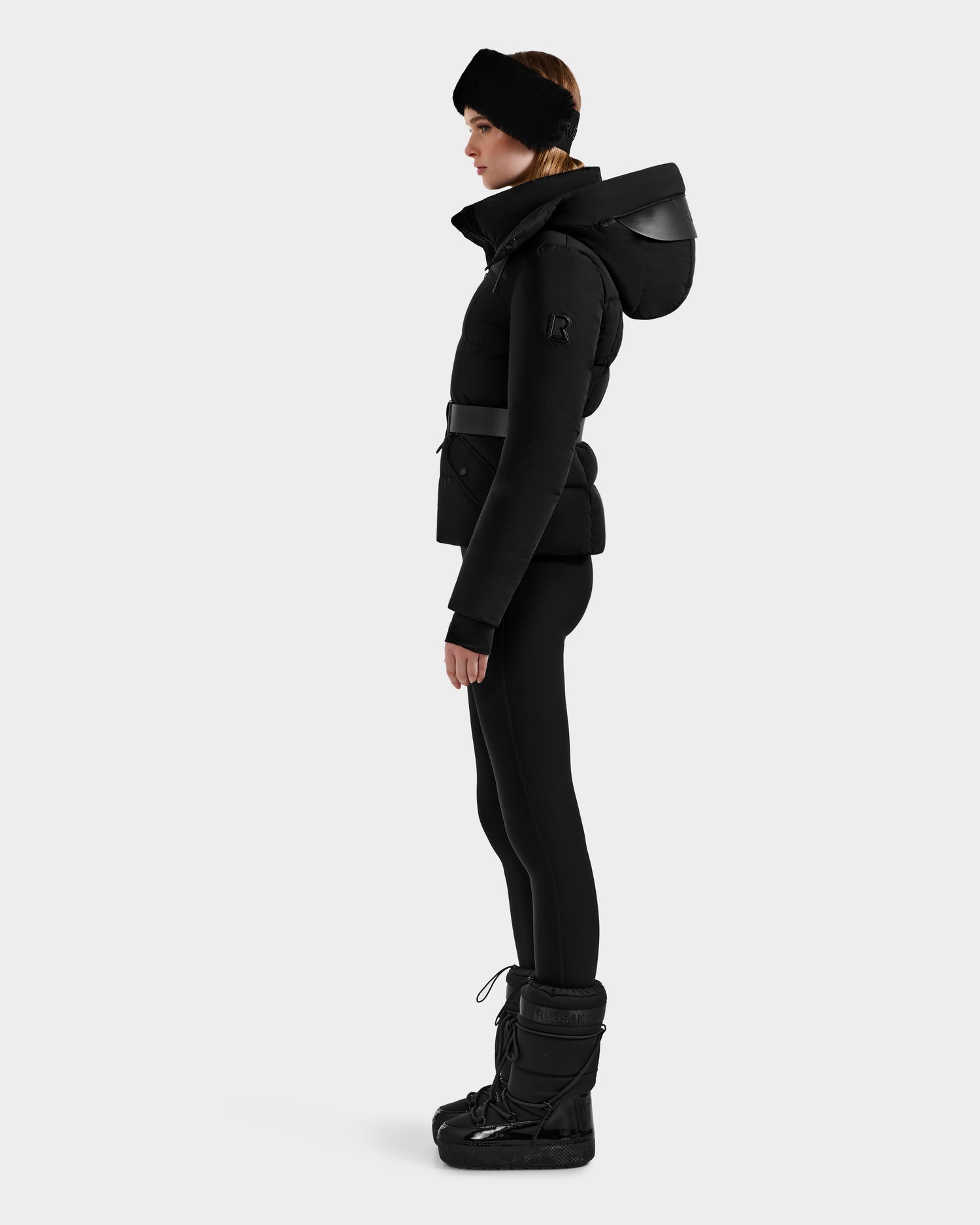 A person stands in profile wearing the Rudsak ADELINE - 8124544 Black women's down puffer jacket with a belt, paired with black leggings, winter boots, and a headband, set against a plain light gray background.