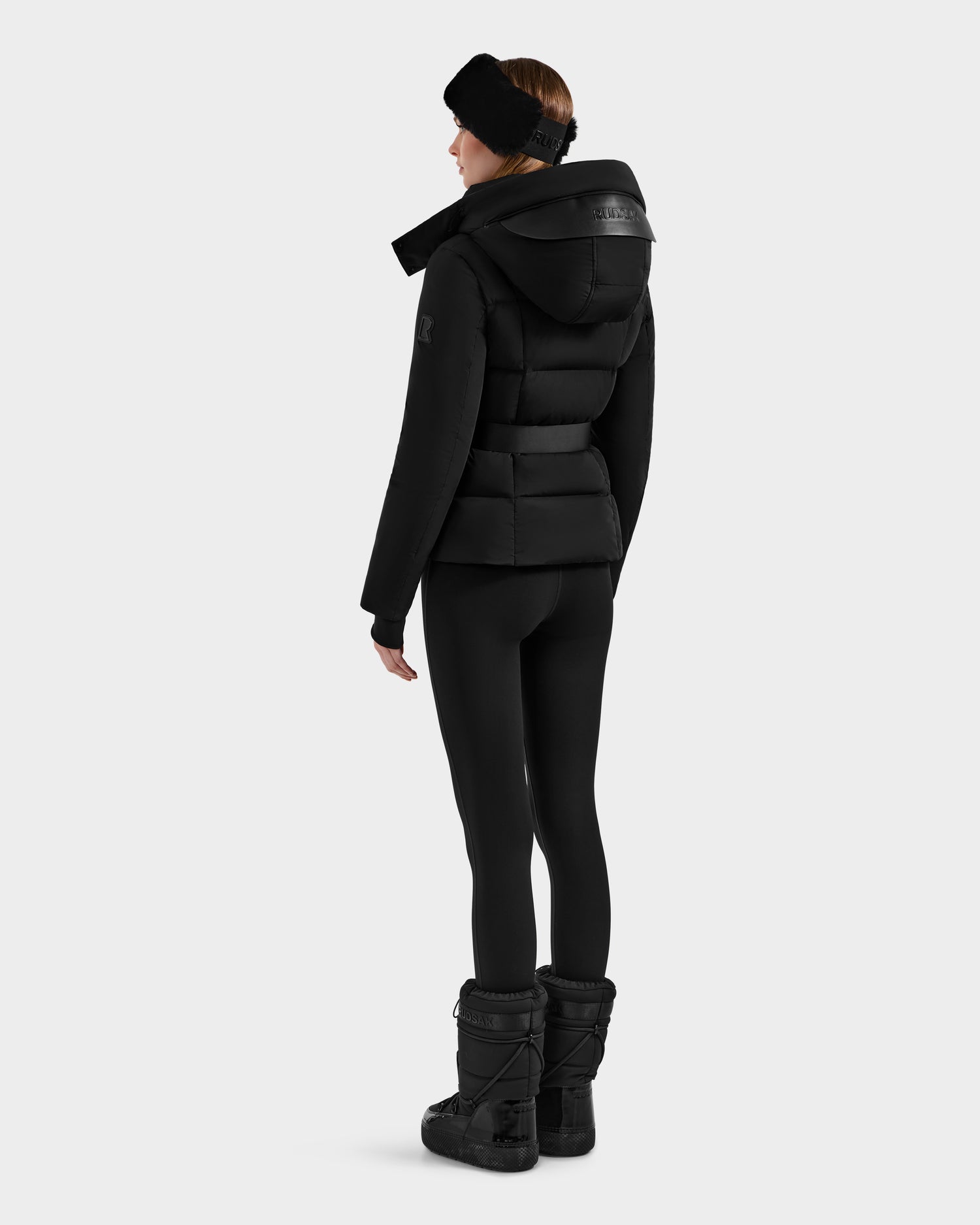 A woman faces away against a light background, wearing the Rudsak ADELINE - 8124544 Black quilted winter jacket with a hood, paired with black leggings, winter boots, and a headband.