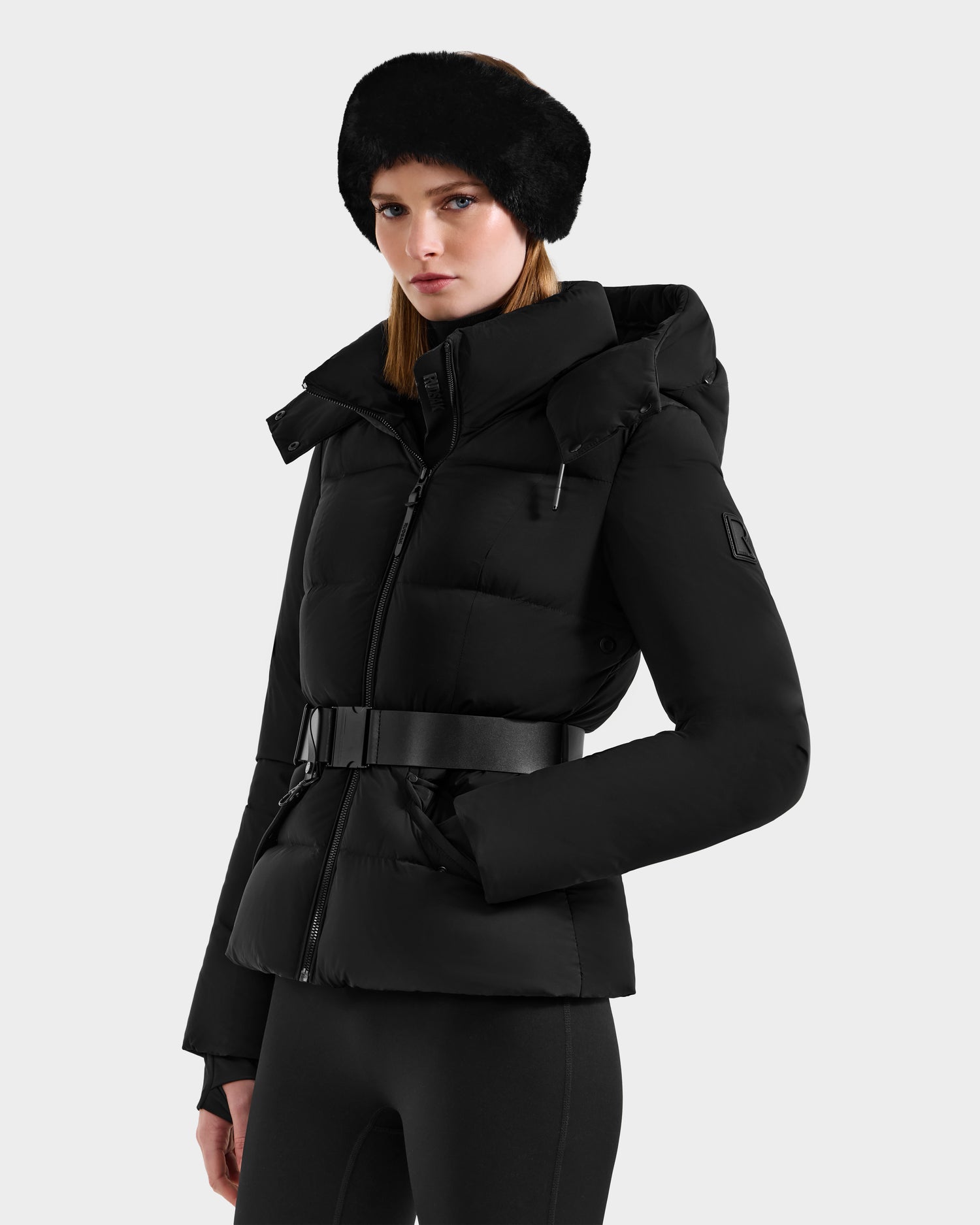 A woman in the Rudsak ADELINE - 8124544 Black quilted winter jacket with a high collar, wide black belt, and fur headband stands against a plain light background, gazing slightly to the side.