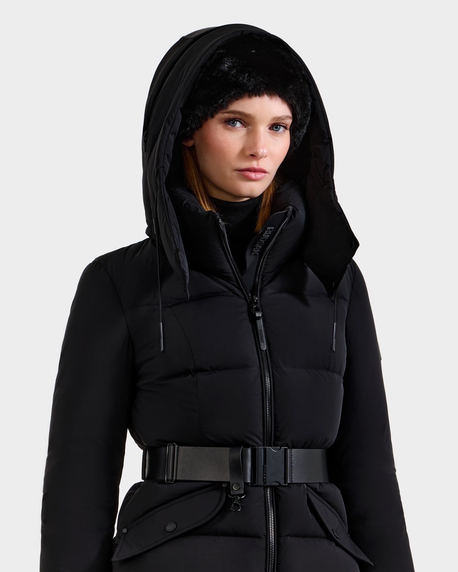 A woman models the Rudsak ADELINE - 8124544 Black quilted winter jacket with a wide belt and a black fur hat, looking forward with a neutral expression against a plain light background.