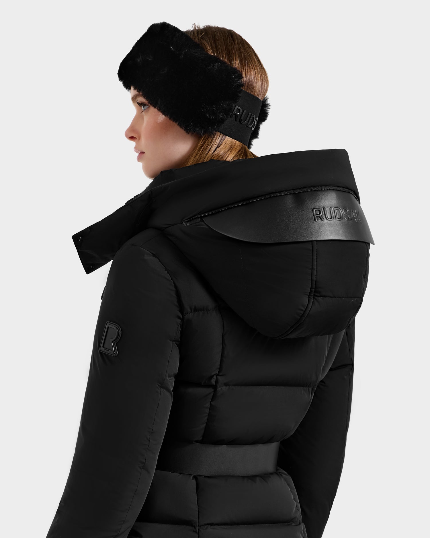 A woman, seen from the back against a plain light background, wears the ADELINE - 8124544 Black down puffer jacket and a black fur headband, both by Rudsak and featuring the brand's logo.