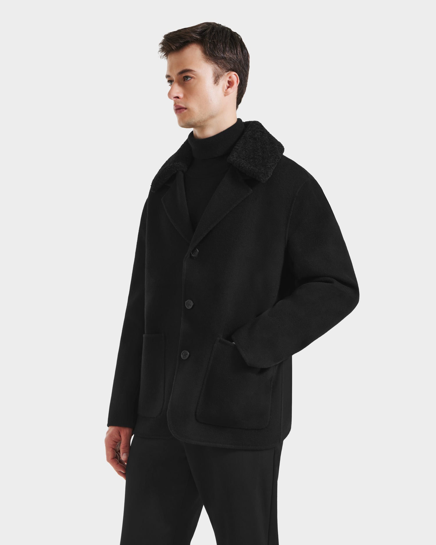 A man wearing the Rudsak AIDEN - 6125558 Black men’s wool-blend coat with large front pockets and a black turtleneck stands against a plain white background.