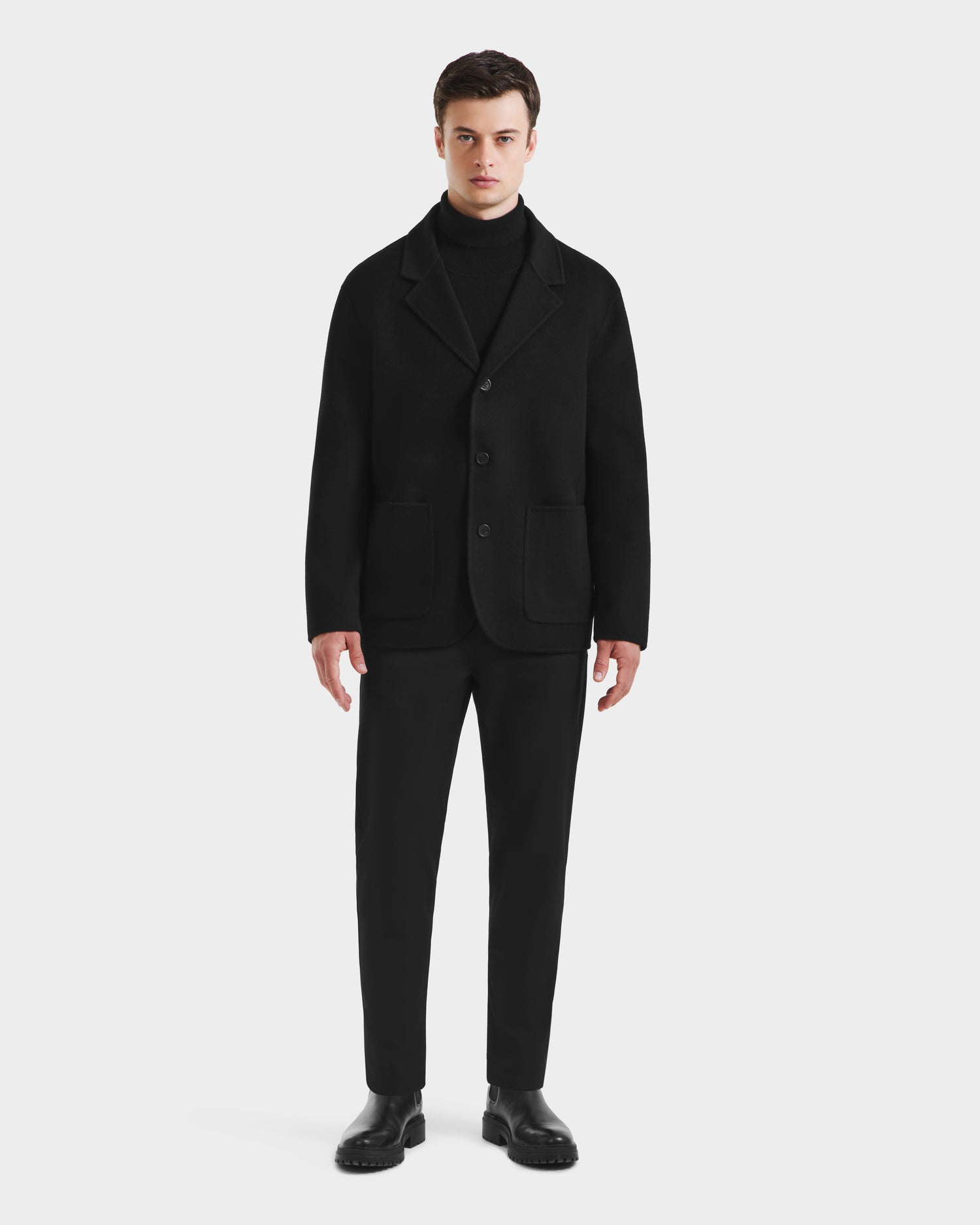 A man faces forward in a stylish outerwear look, wearing the Rudsak AIDEN - 6125558 Black men’s wool-blend coat with a removable faux shearling collar, paired with black pants and dress shoes against a plain white background.
