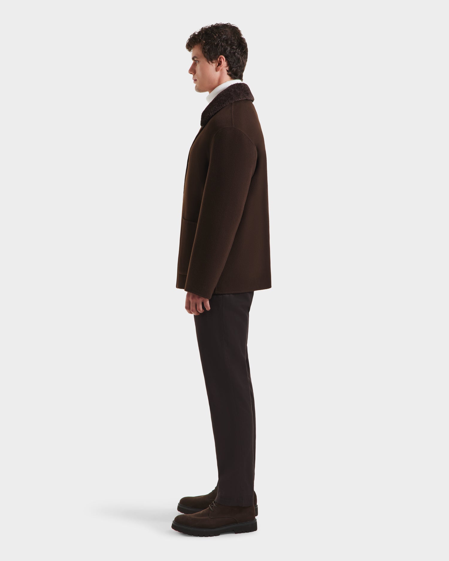 A man stands in profile facing left, wearing the Rudsak AIDEN - 6125558 Espresso men’s wool-blend coat with a removable shearling collar, paired with dark pants and shoes against a plain white background.