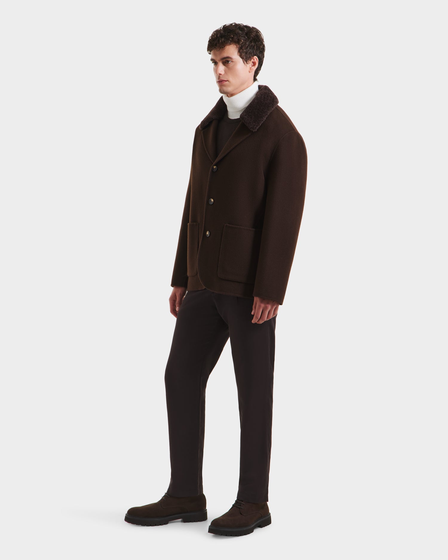 A man stands against a plain background wearing the Rudsak AIDEN - 6125558 Espresso coat—a dark brown men’s wool-blend piece with large front pockets—paired with a white turtleneck, dark pants, and brown shoes. He faces slightly to the side with a neutral expression.