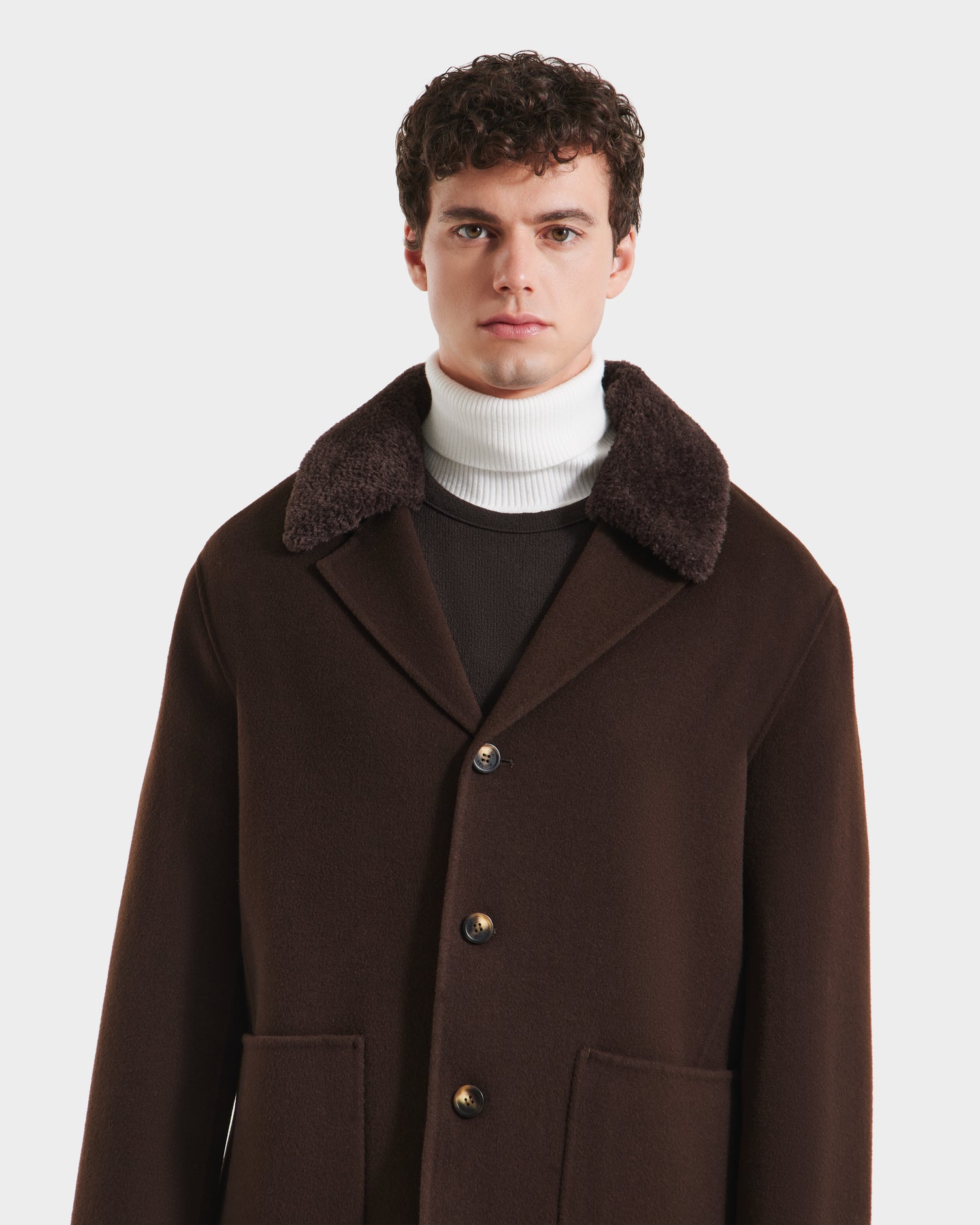 A man with short curly hair models the Rudsak AIDEN - 6125558 Espresso wool-blend coat, featuring a wide removable shearling collar, layered over a white turtleneck against a plain white background.